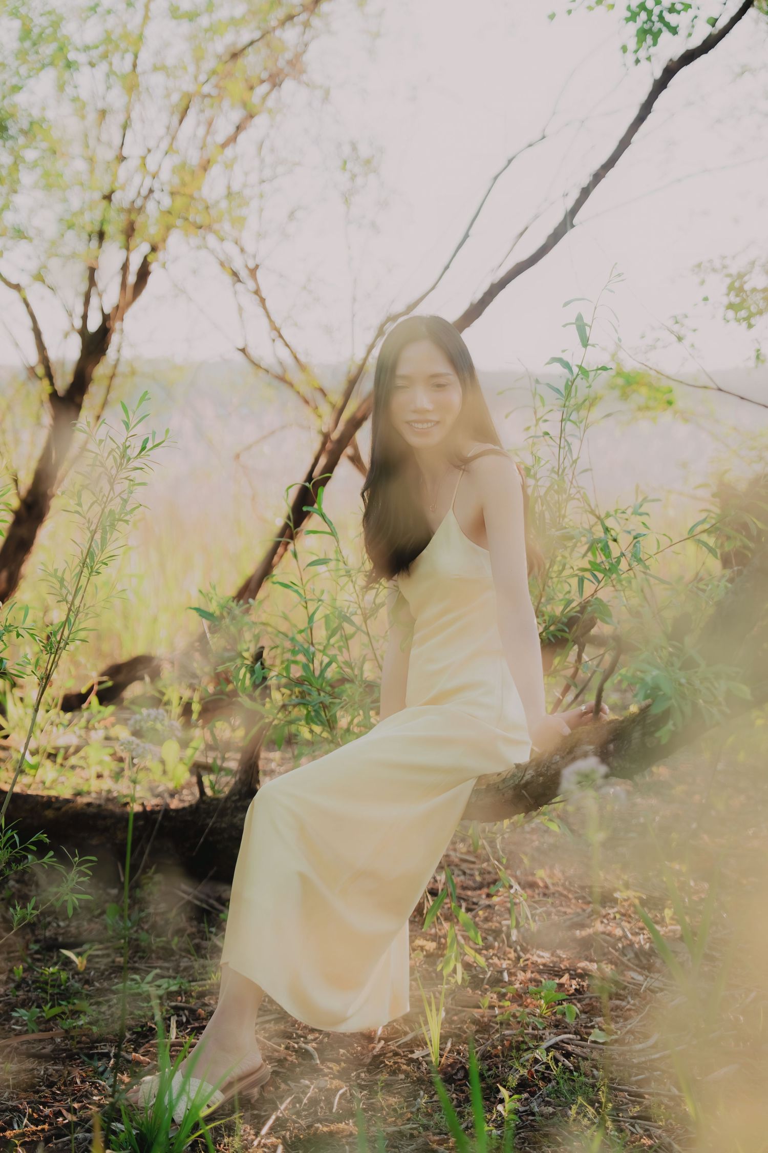 Young woman in cream slip dress sits among wildflowers and trees in a dreamy, sun-dappled forest setting.
