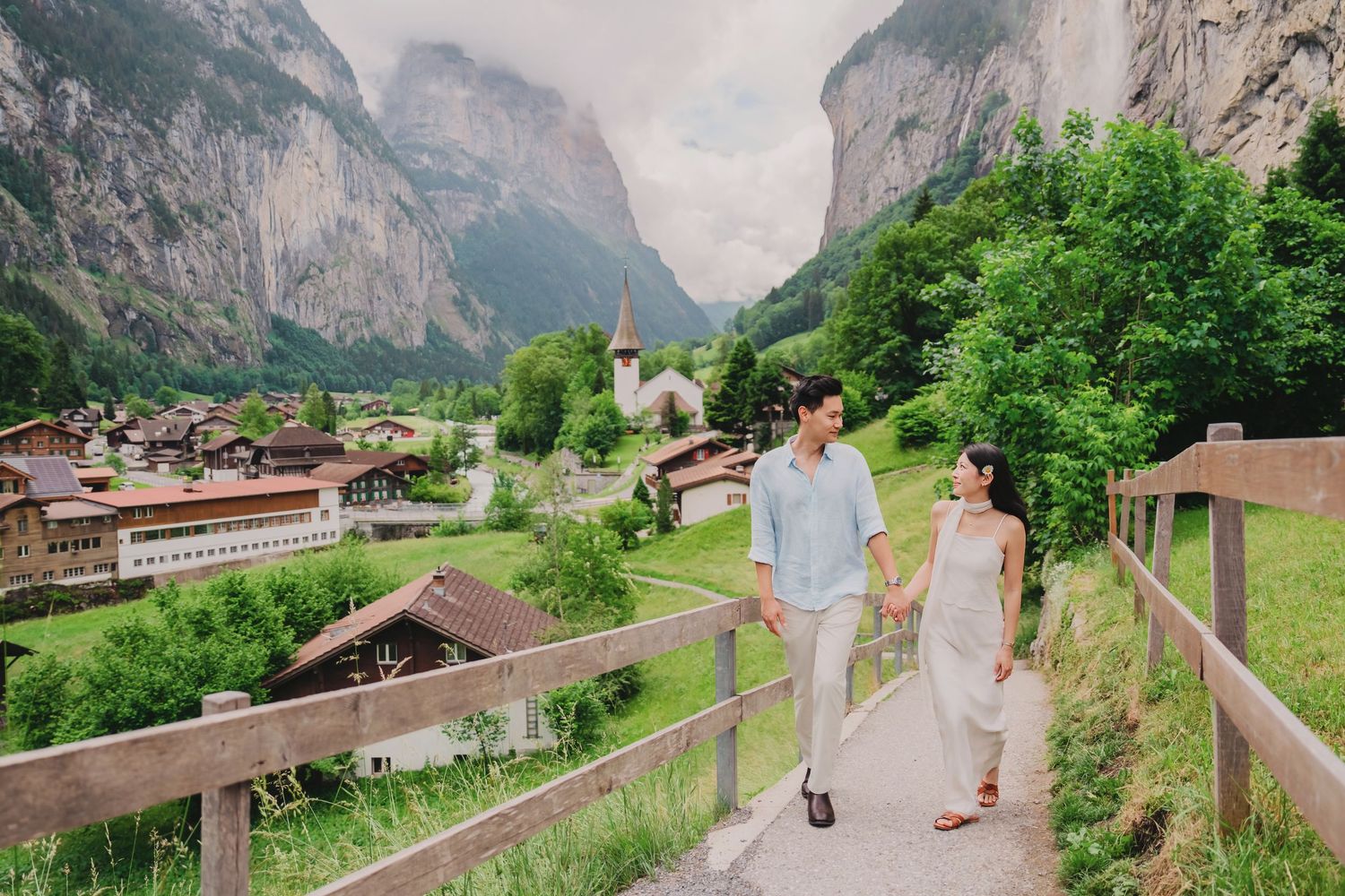 Couple walking hand in hand on a scenic path in Lauterbrunnen, Switzerland, with alpine village and misty cliffs backdrop.