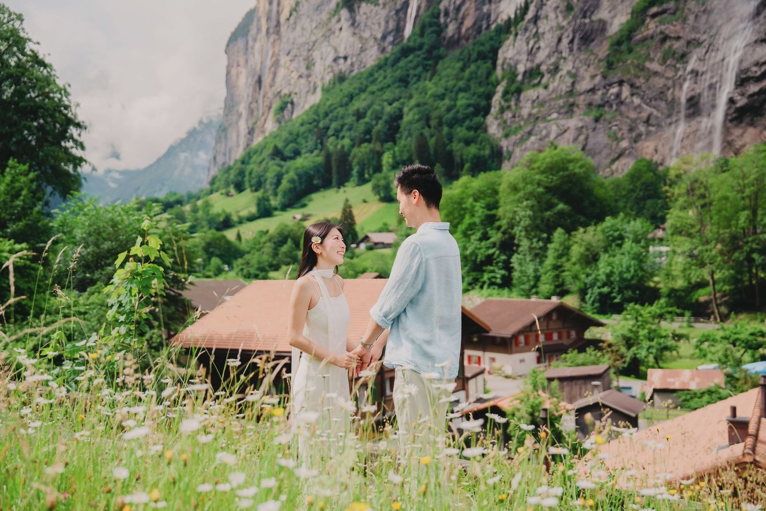 Couple holds hands facing each other in a wildflower meadow with Swiss chalets and alpine cliffs behind them.