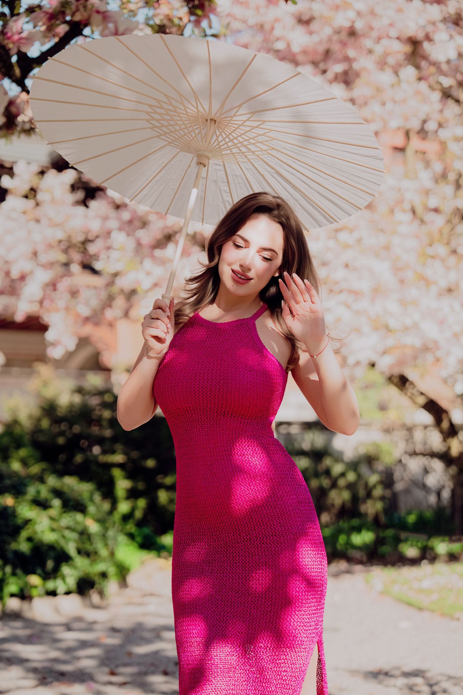 Woman in pink knit dress holding white parasol near cherry blossoms.