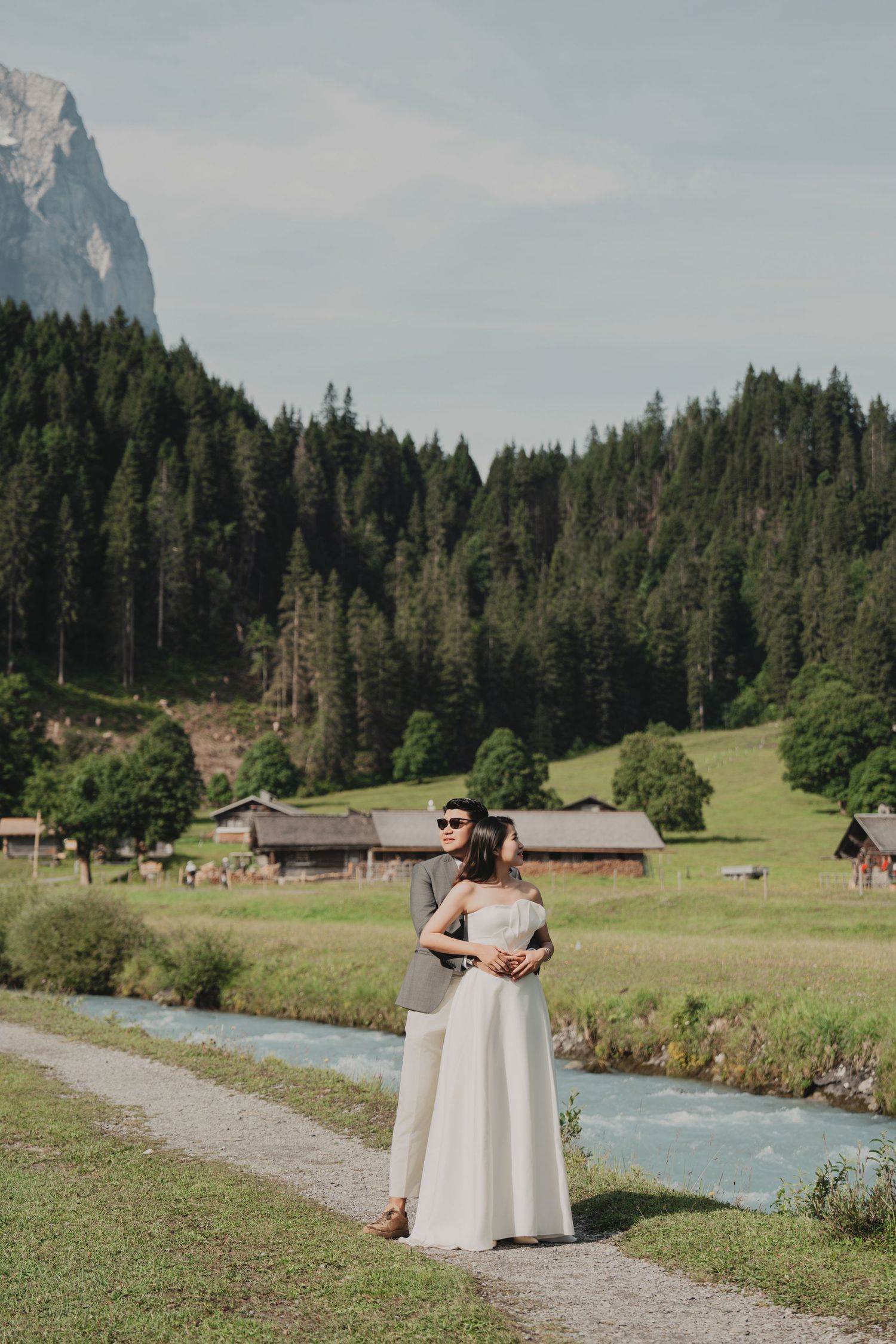 Groom embracing bride from behind on a riverside path with wooden chalets and forest scenery.