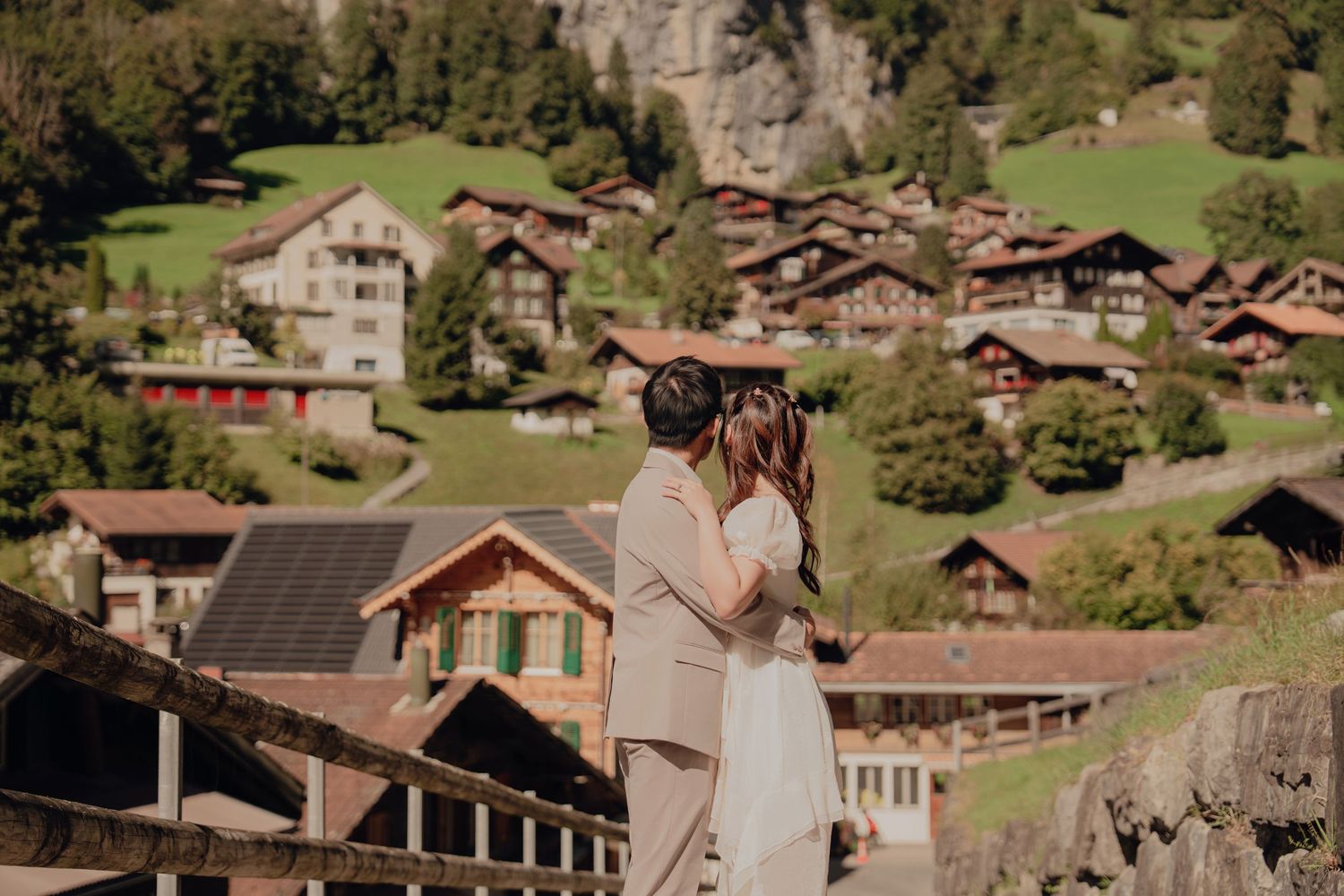 Romantic couple sharing a tender moment on a bridge overlooking a picturesque Swiss mountain village.