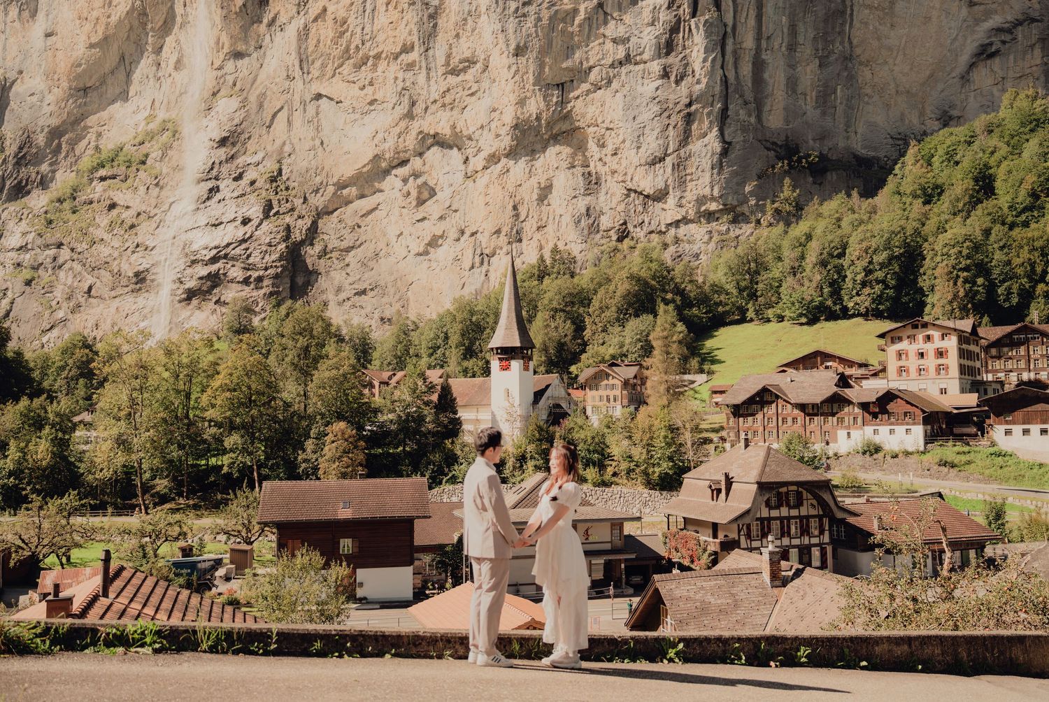 Couple holding hands facing a picturesque Swiss village with a church and rocky cliffs.