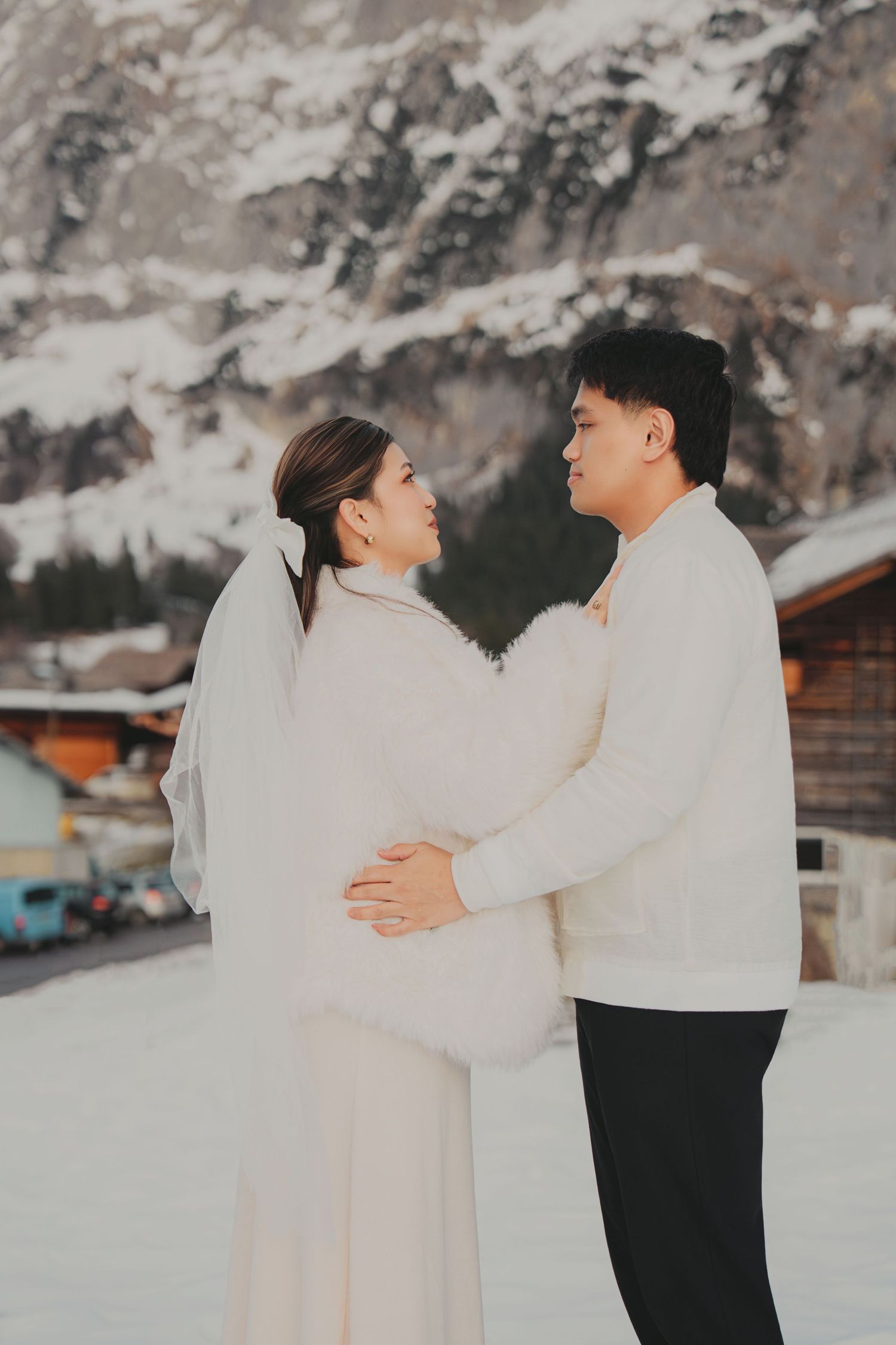 Couple in white outfits sharing a tender moment embracing in a snowy alpine village landscape.