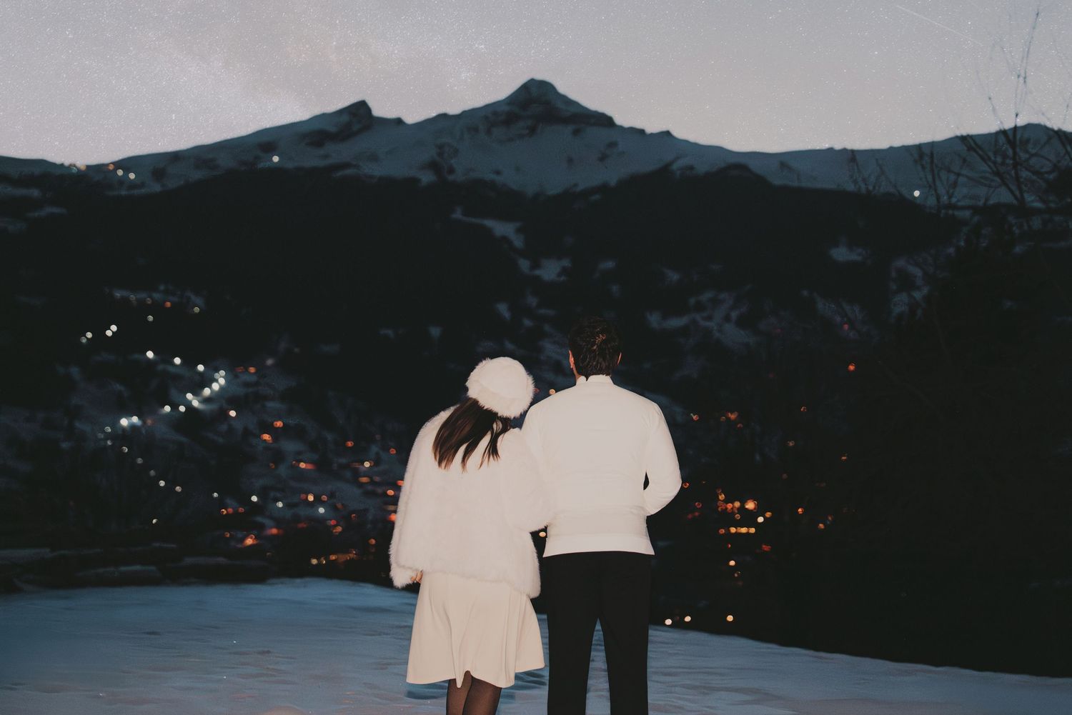 Couple in white outfits viewing snow-covered mountains at twilight with valley lights.
