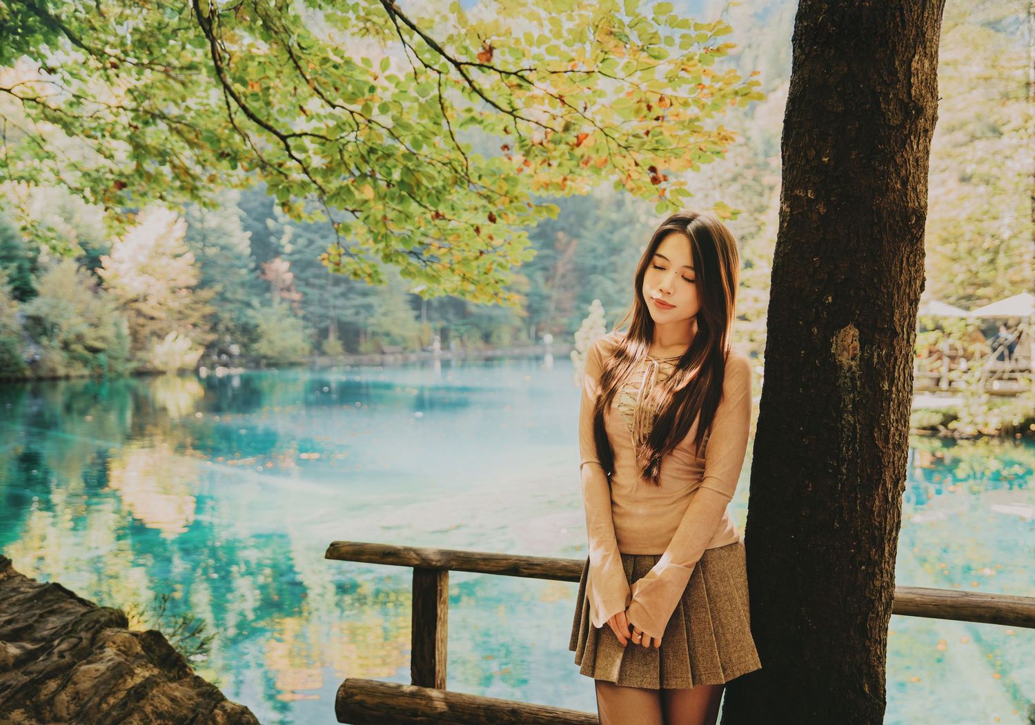 Young Asian woman in beige sweater and skirt stands by turquoise lake under autumn tree branches.