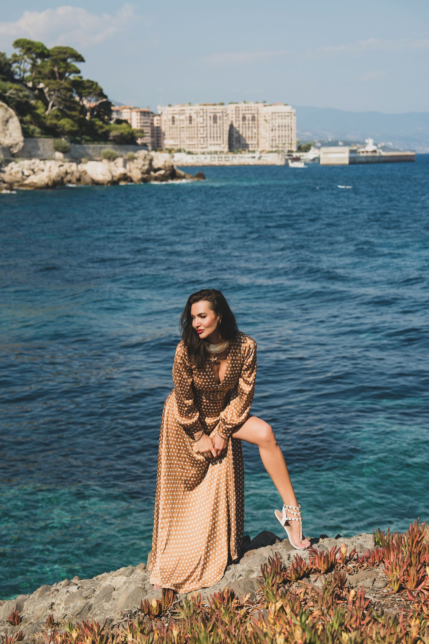 Brunette woman in gold polka dot long dress sitting on coastal rocks with blue sea backdrop.