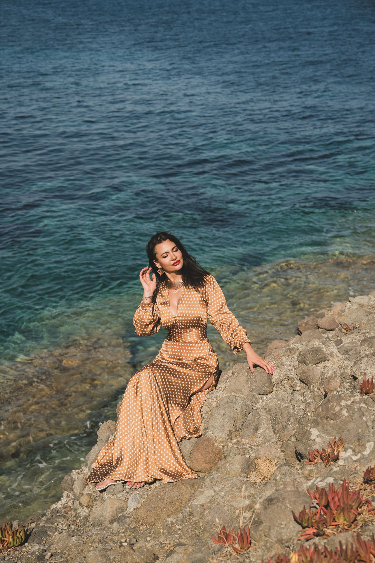 Woman in elegant polka dot dress sits on rocky shore beside turquoise Mediterranean waters.