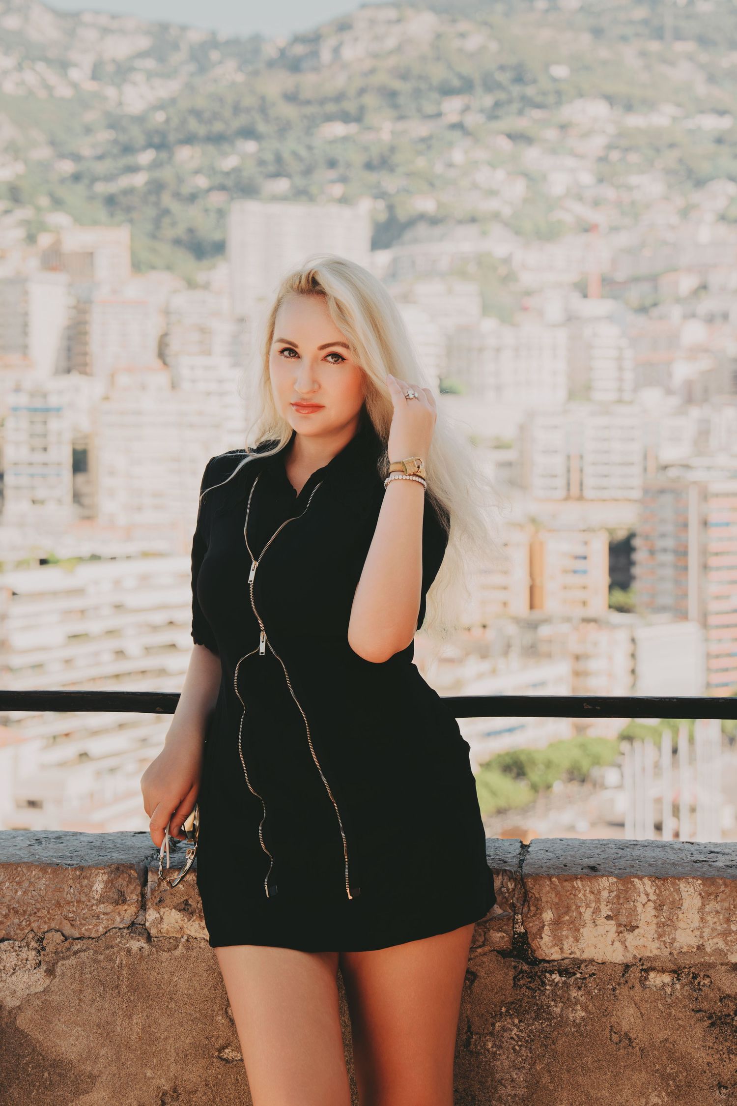 Blonde woman in black zip dress posing on balcony with Monaco cityscape behind her.