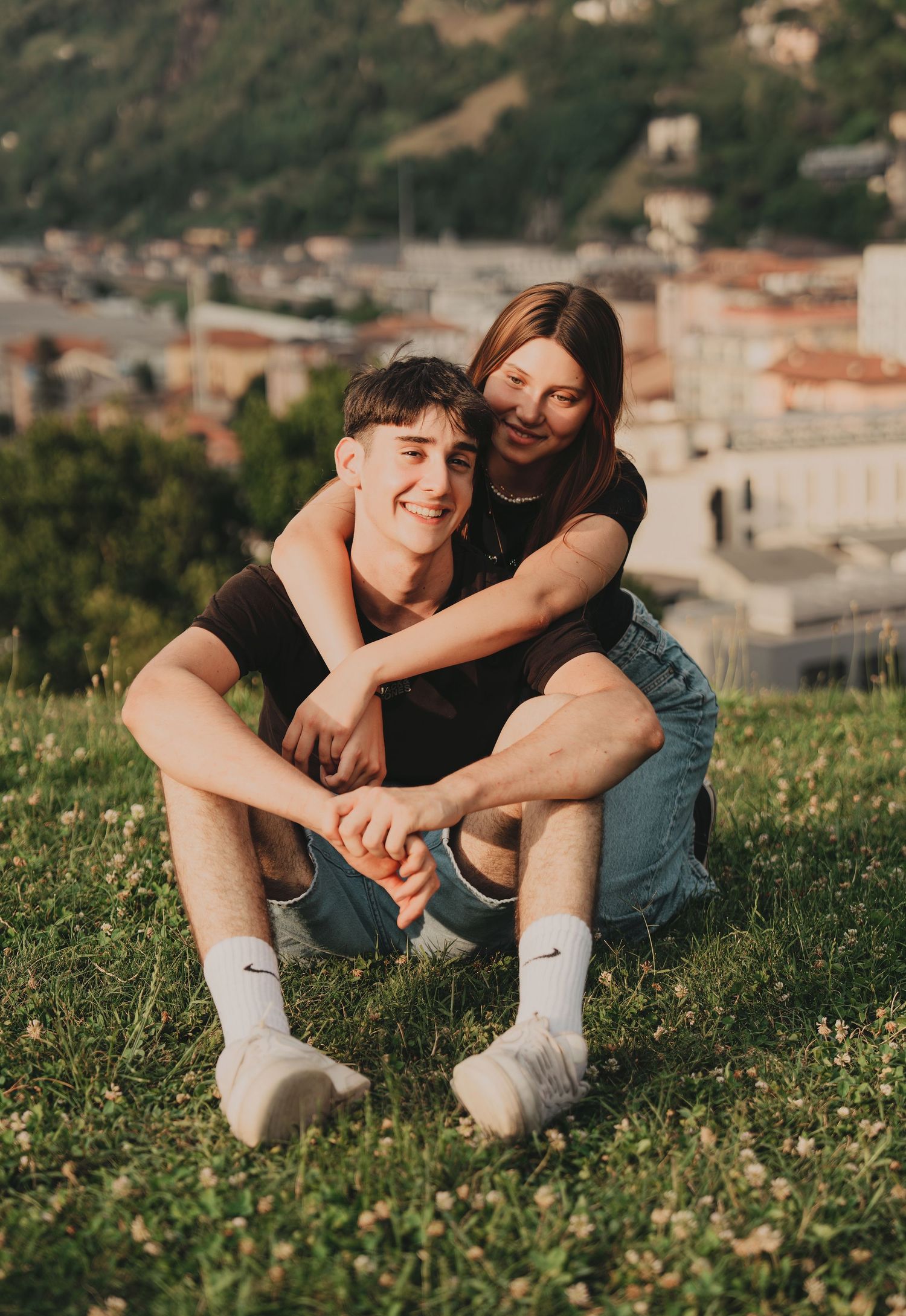 Happy young couple embracing on green grass with scenic town backdrop at sunset.