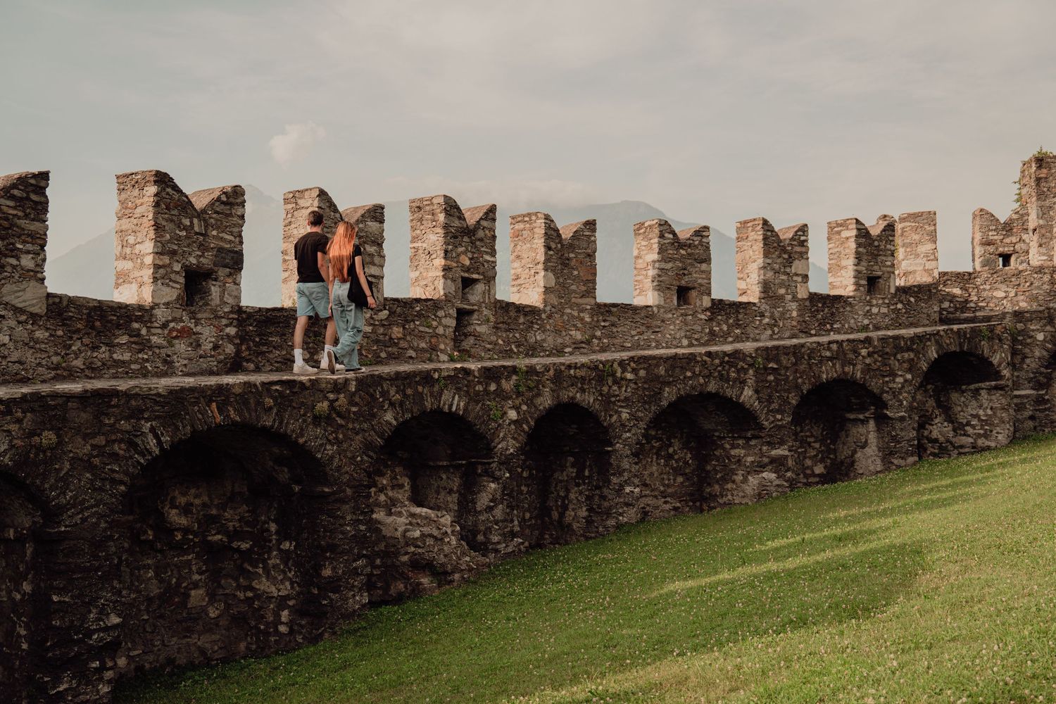 A couple strolling along the top of a historic medieval castle wall with arched buttresses below.