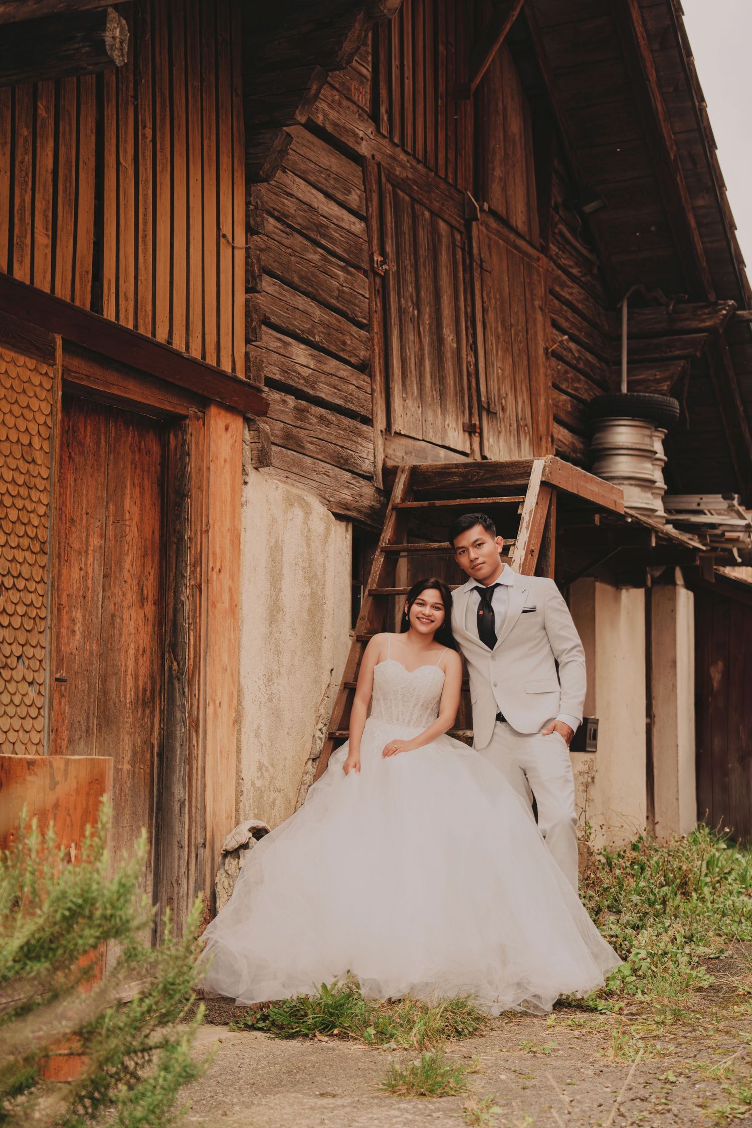 Bride in white ballgown and groom in ivory suit pose together beside a rustic wooden barn.