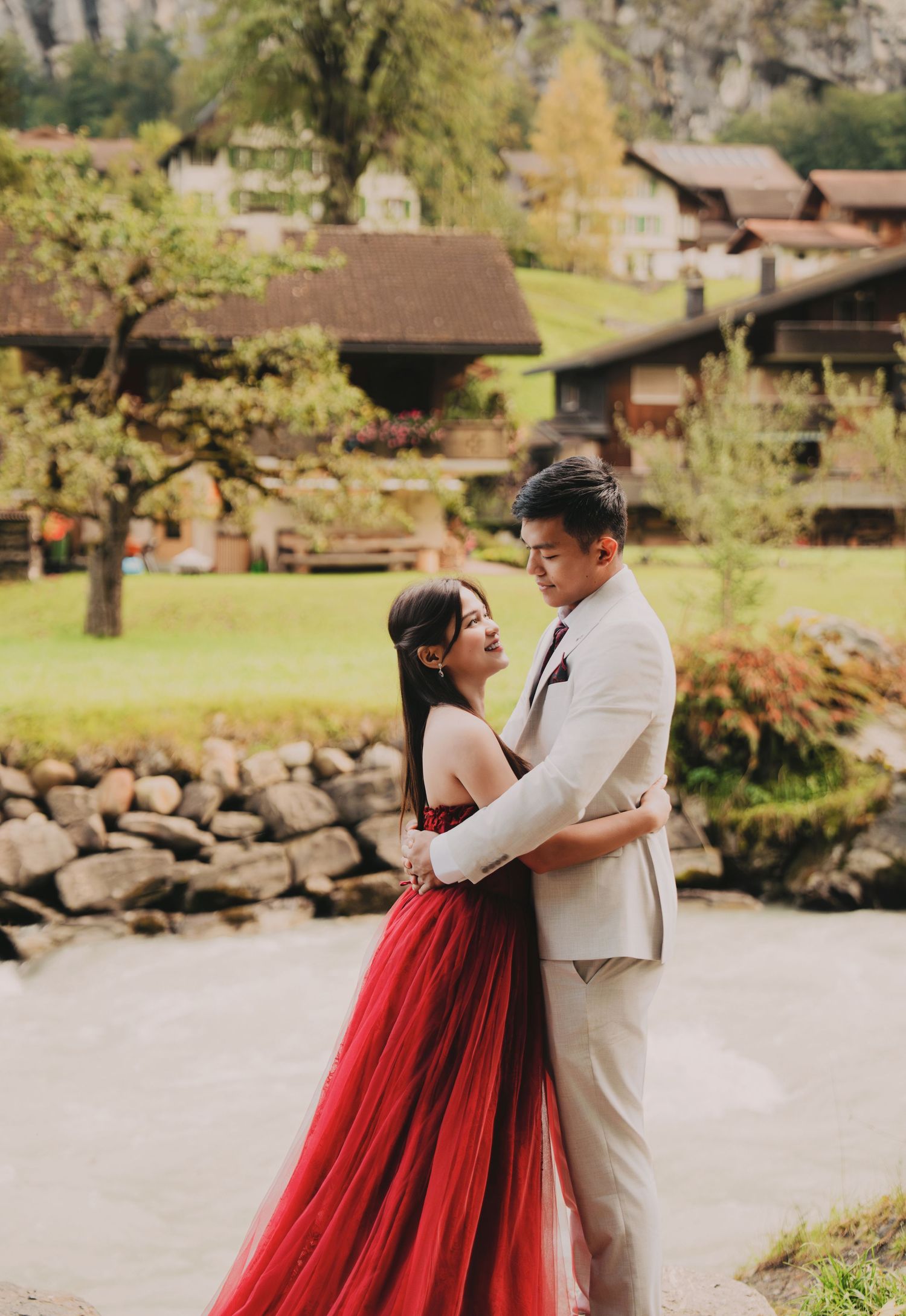 Asian couple embracing tenderly in red bridal gown by riverside with Swiss chalet village backdrop.