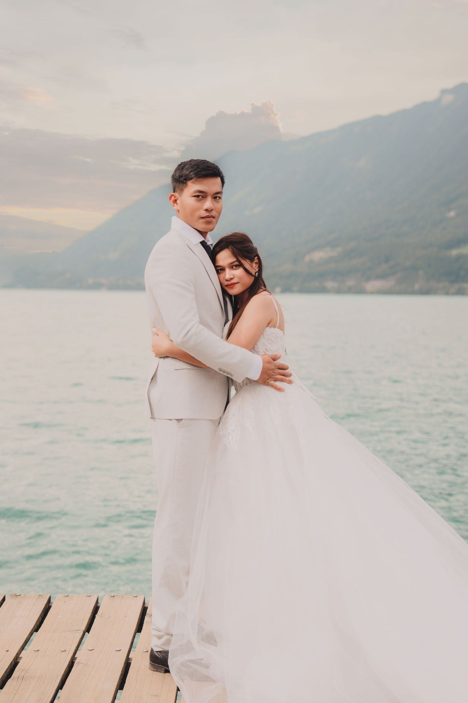 Wedding couple embracing on lakeside pier with stunning mountain scenery and cloudy sky behind them.