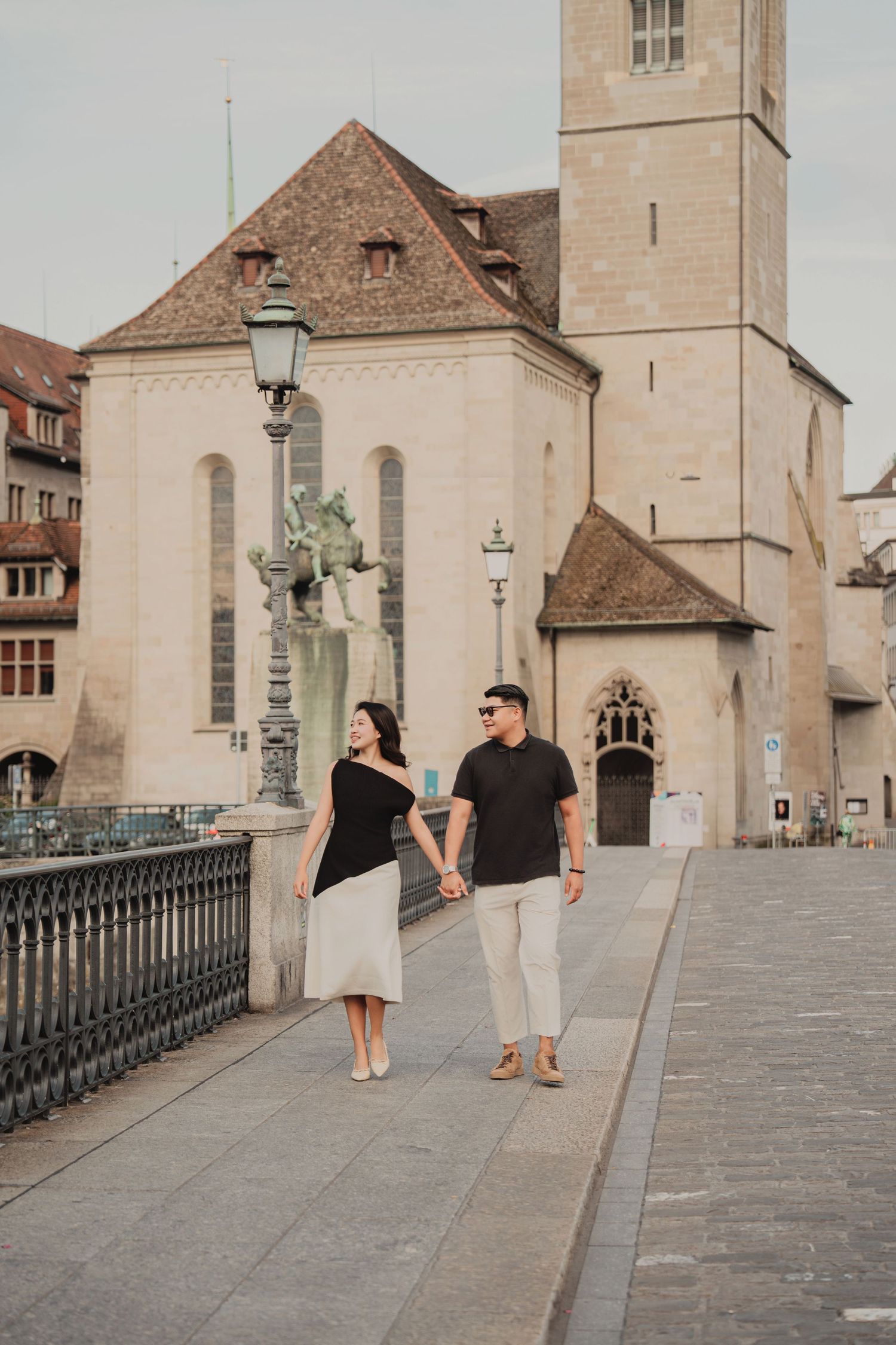 Couple holding hands strolling along a historic Zurich bridge with a medieval church in background.
