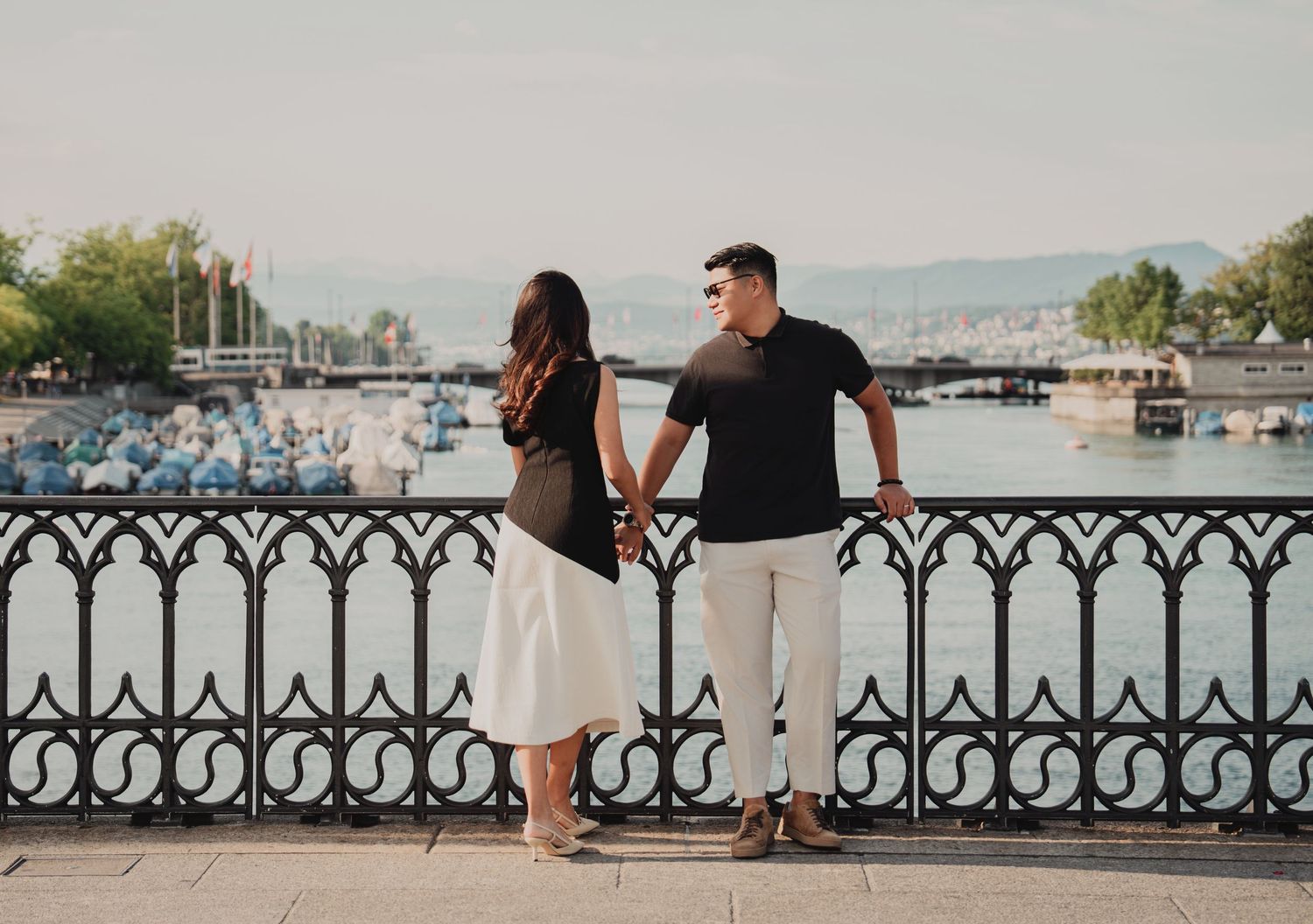 Couple holding hands facing Zurich harbor with boats and city skyline in warm sunlight.