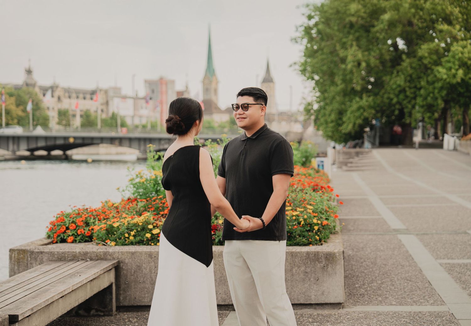 Asian couple facing each other by Zurich's riverside promenade with blooming flowers and church towers.