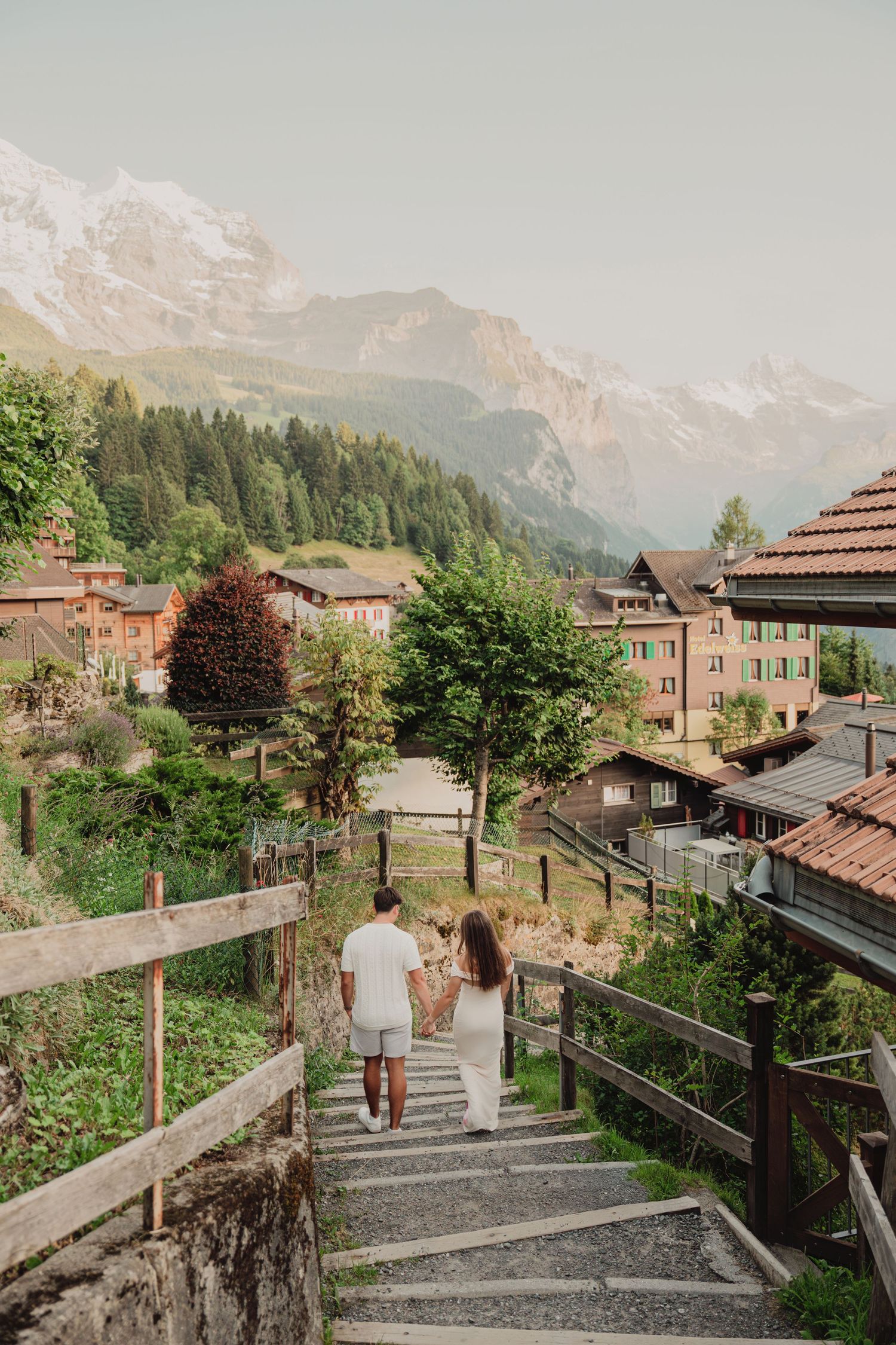 Couple strolling down rustic stairway through charming Alpine village surrounded by lush greenery and mountains.