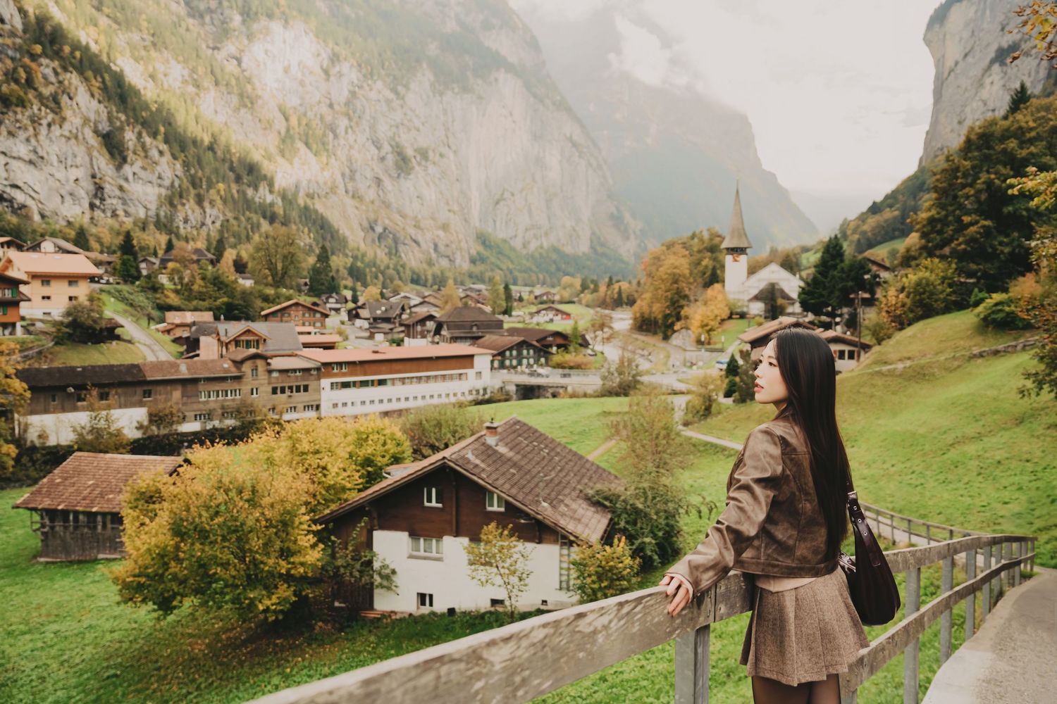 Woman admiring a breathtaking Swiss alpine village with colorful autumn trees and misty mountains.