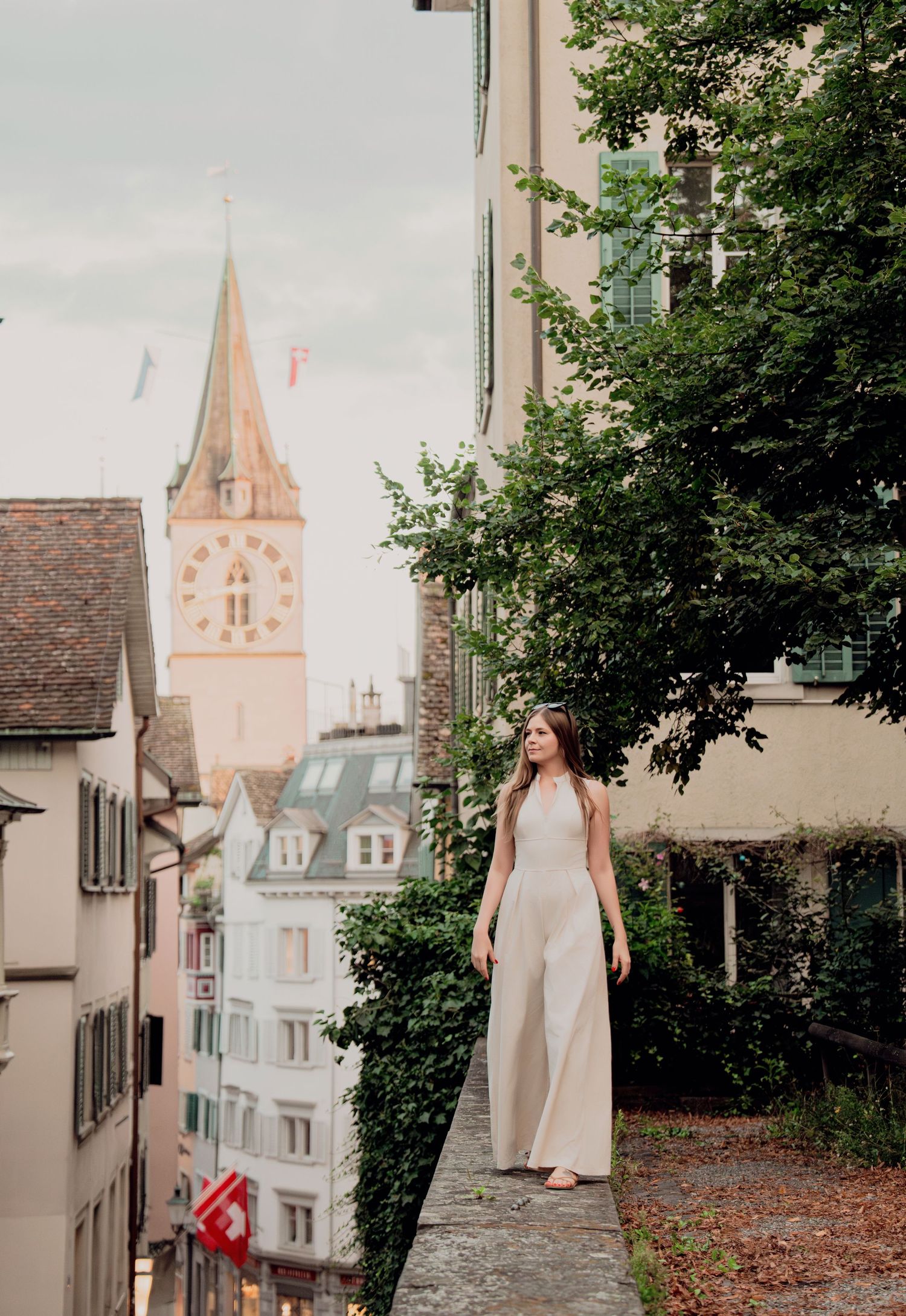 Woman in white jumpsuit walking on historic European stone path with tall church tower and Swiss flags nearby.