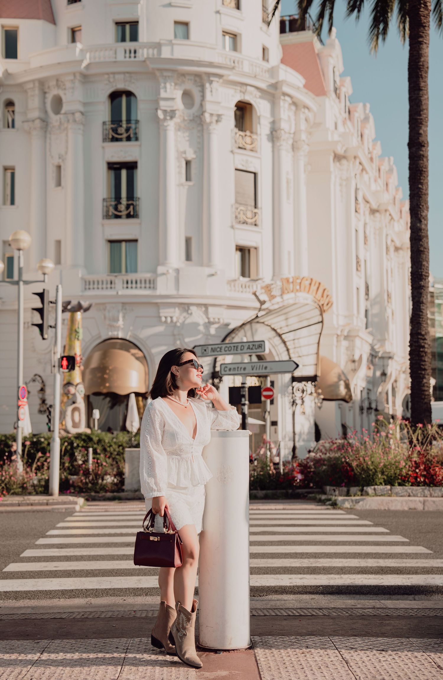 Elegant woman in white outfit holding burgundy bag near luxury hotel in Nice, France.
