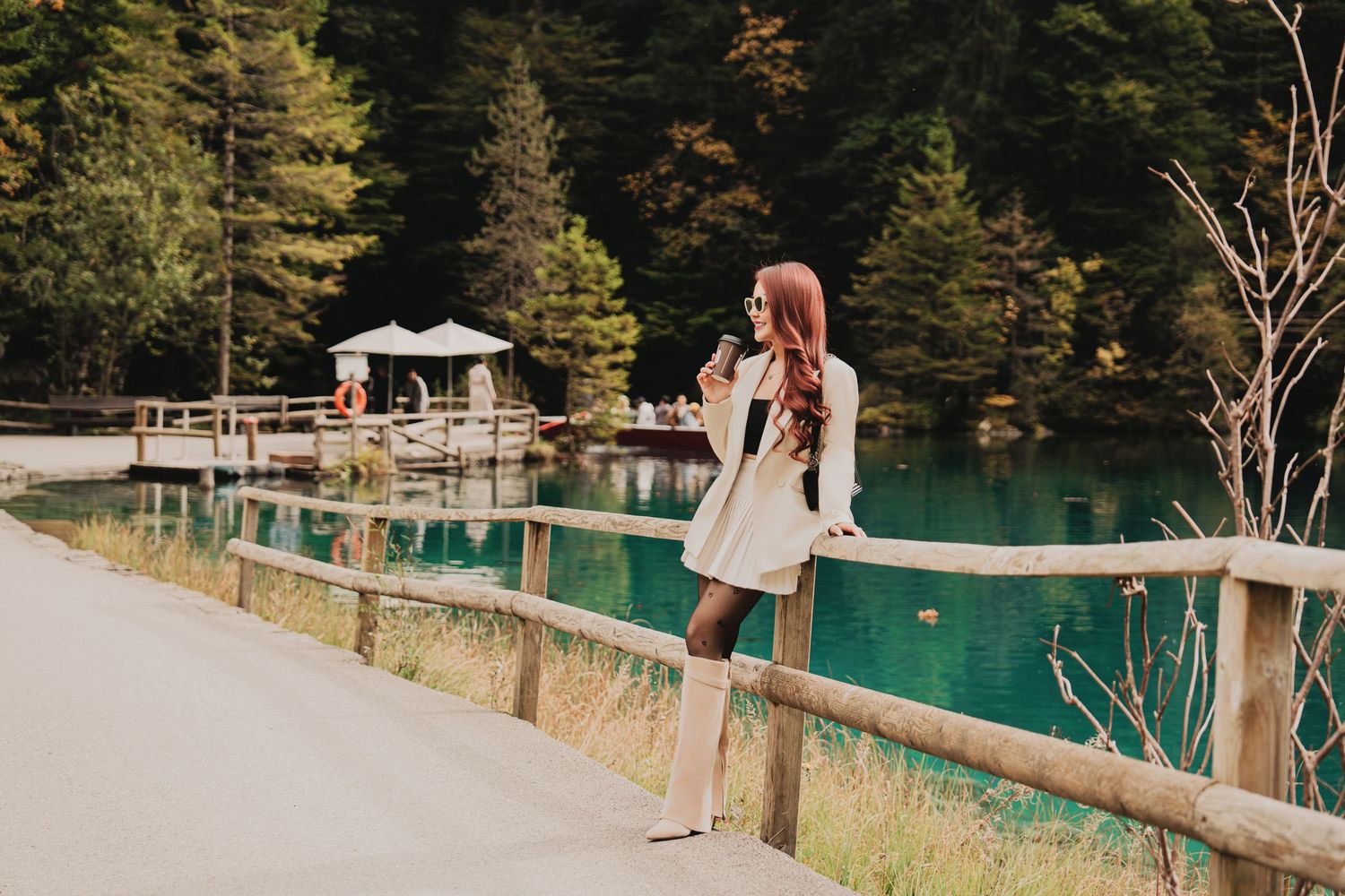 Red-haired woman in white dress photographing scenic turquoise alpine lake from wooden walkway.