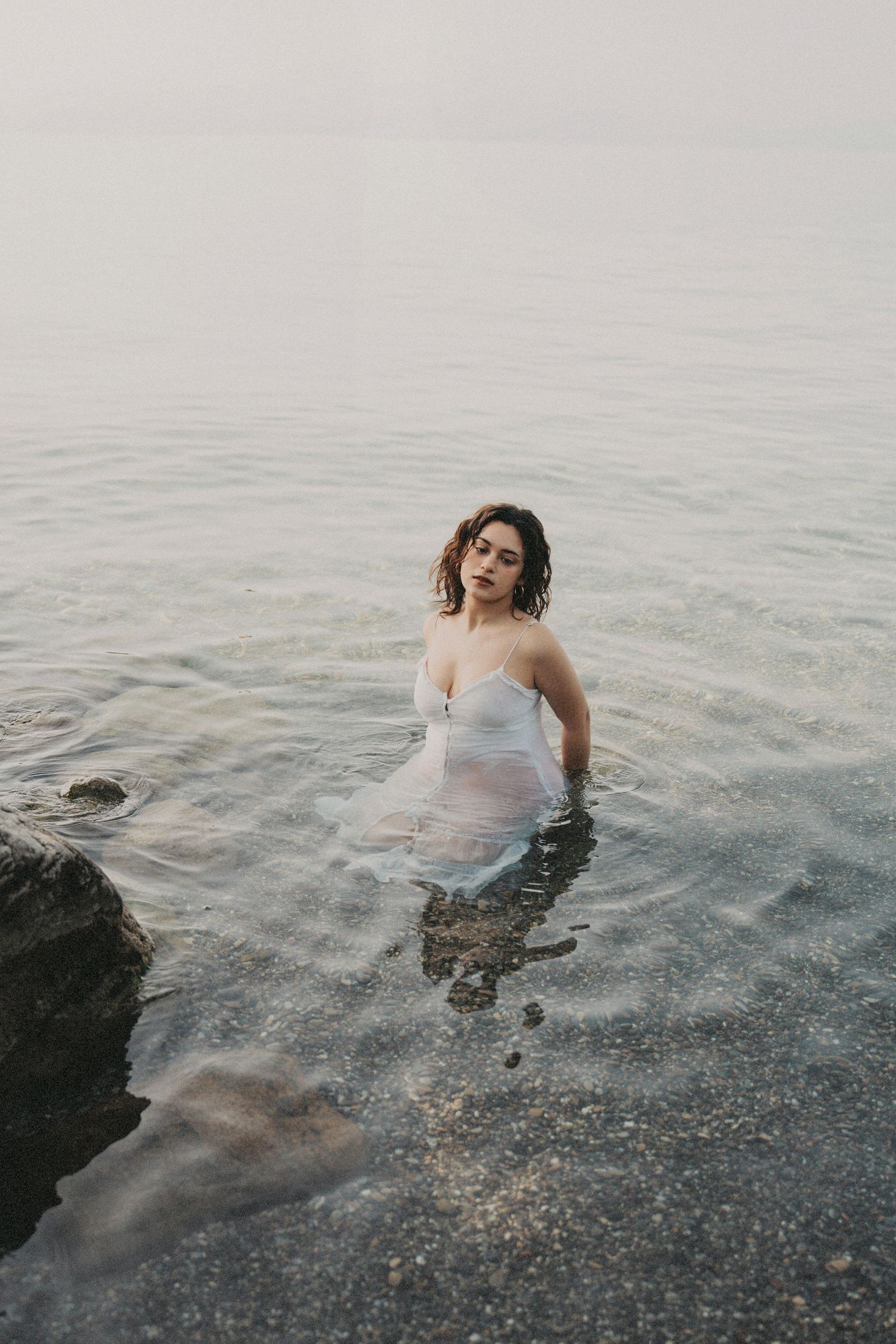 Young woman in wet white spaghetti-strap dress stands waist-deep in calm misty ocean water near rocks.
