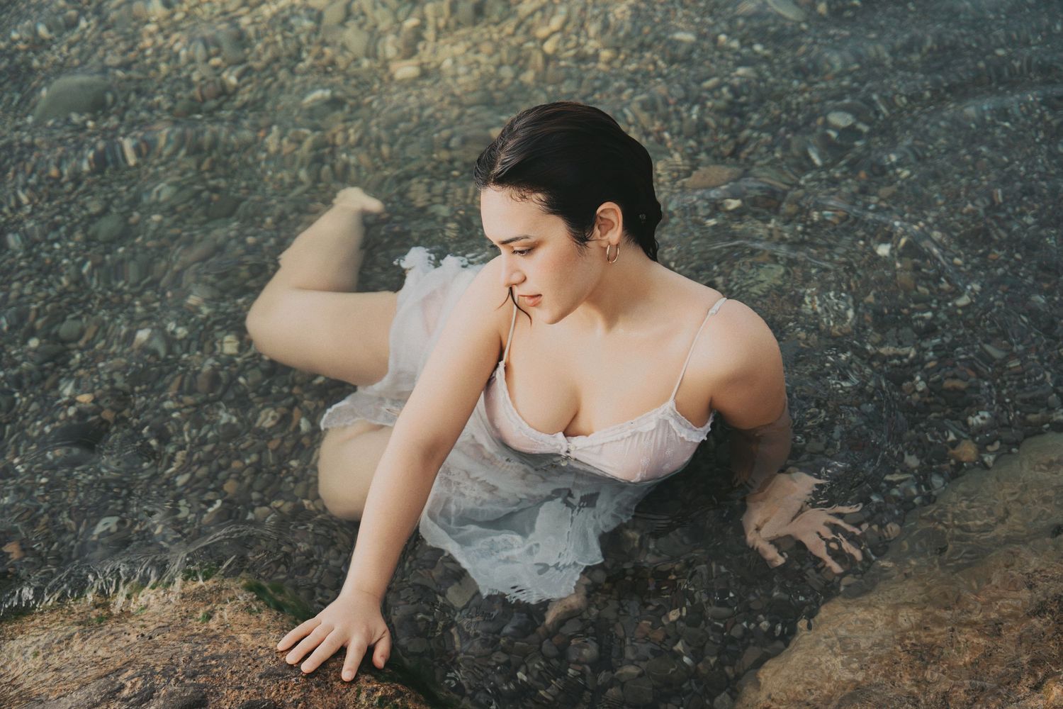 Woman in wet white dress sitting in shallow rocky water, looking to the side in natural light.
