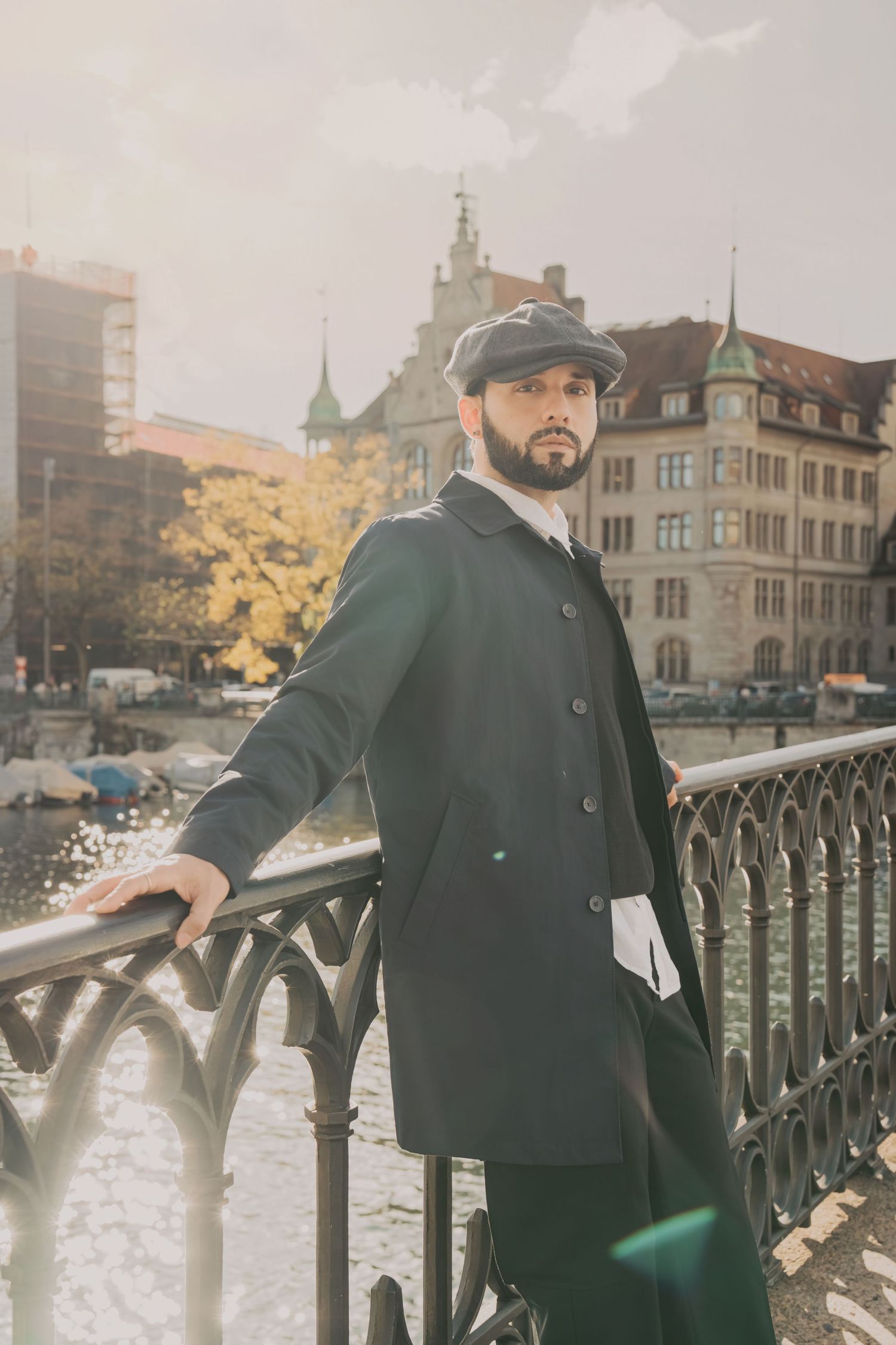 Fashionable man in dark double-breasted coat and flat cap posing on ornate bridge railing in European city.