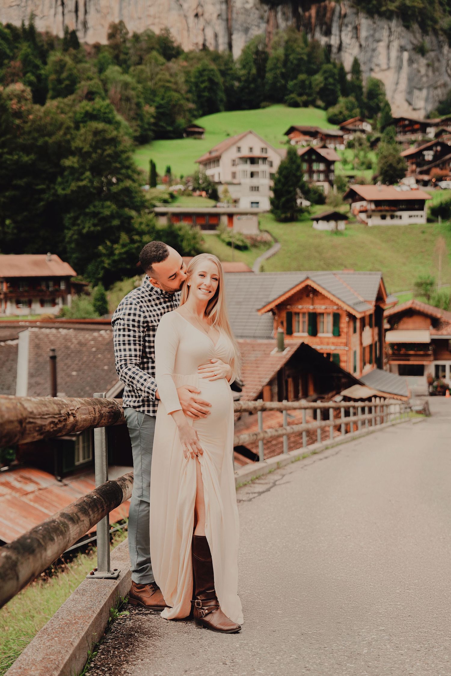 Couple posing on bridge with man embracing pregnant woman in pink dress, Alpine village backdrop.