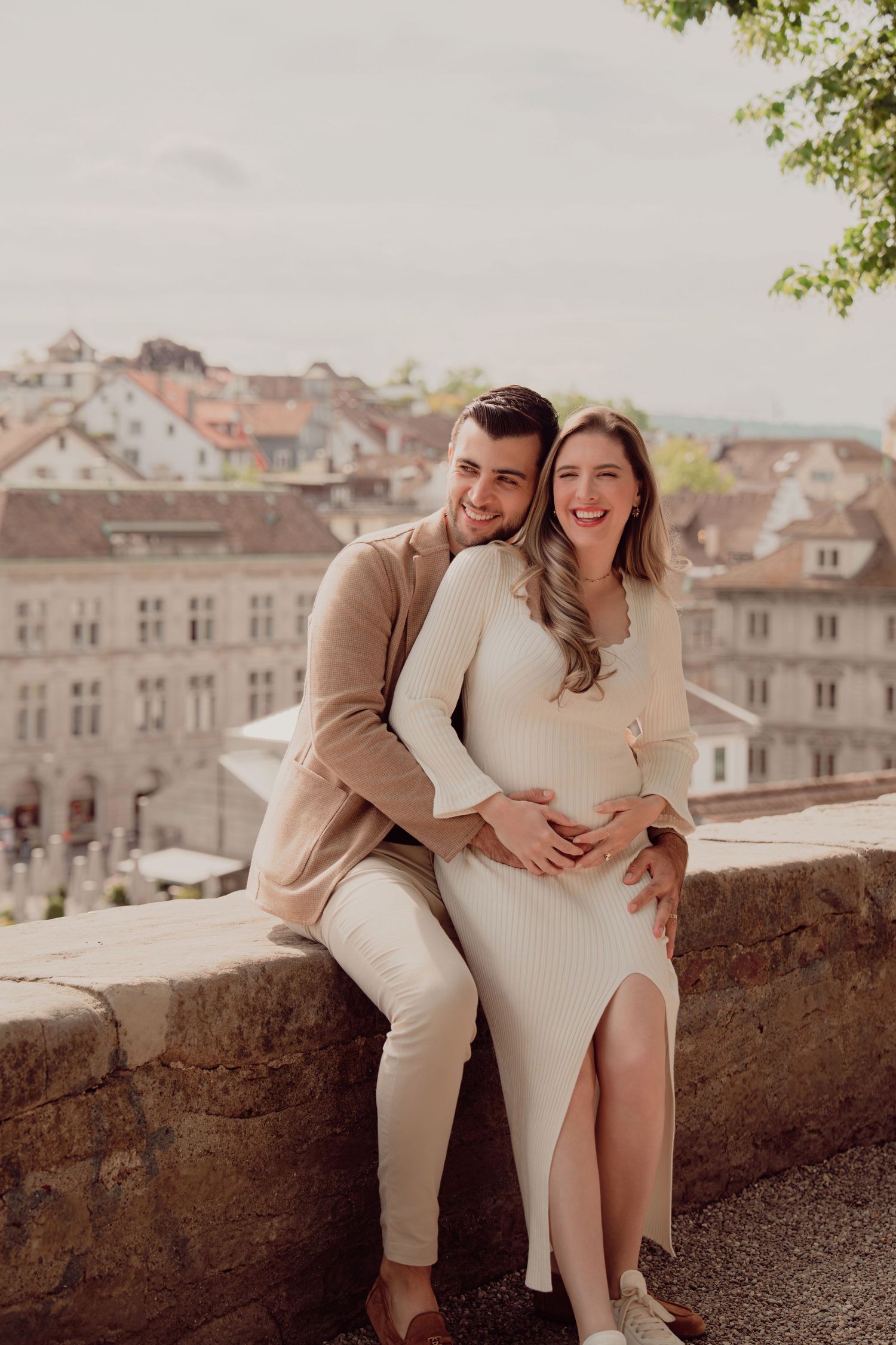 Happy expecting couple sits on stone wall smiling, hands on baby bump, European city in background.