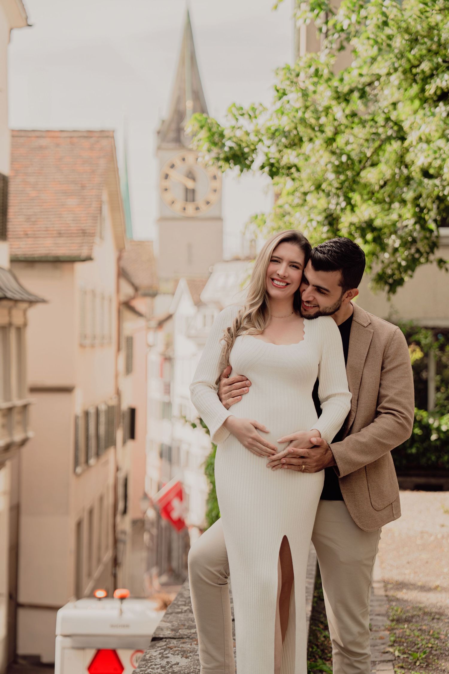 Smiling expectant couple cuddling on a scenic European street, man in tan blazer holding woman's baby bump.