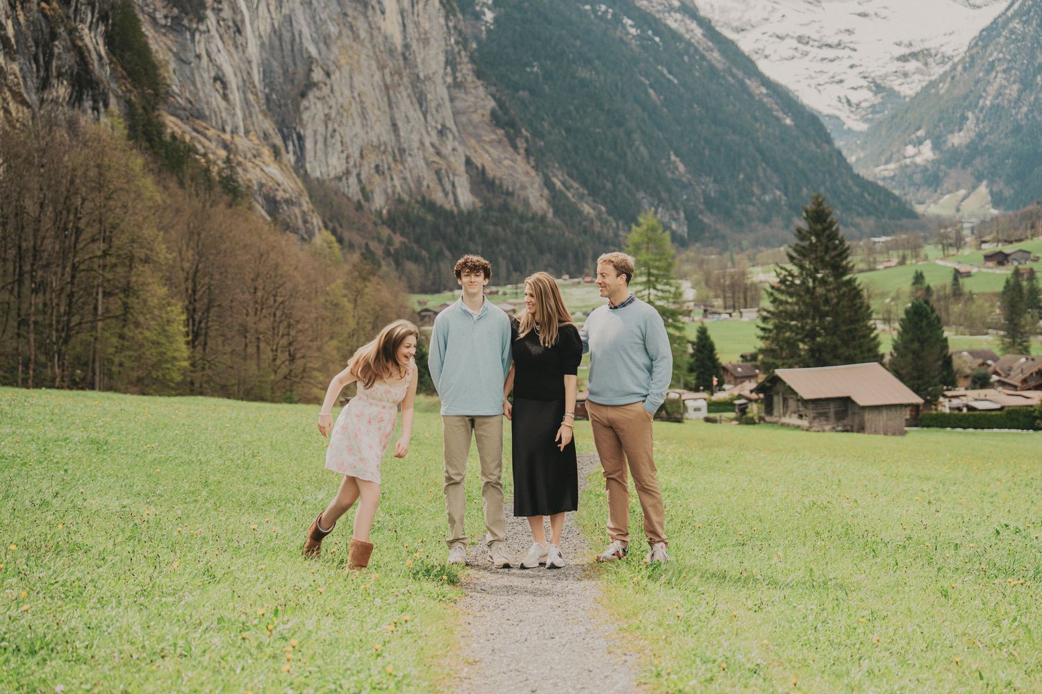 Family laughing together on scenic mountain trail in Swiss Alps with green valley and peaks behind.