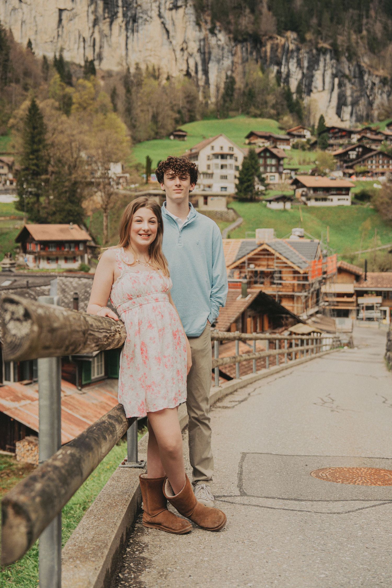 Young couple posing on a bridge in a Swiss alpine village with mountain cliffs and chalets in background.