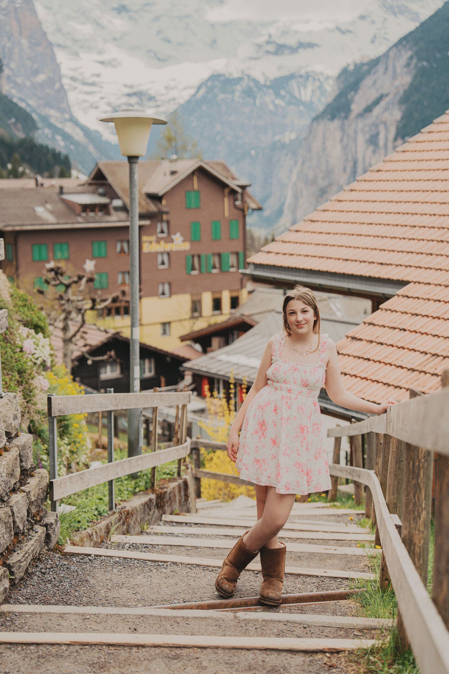 Young woman in pink floral dress and brown boots posing on stairs in charming Swiss mountain village.