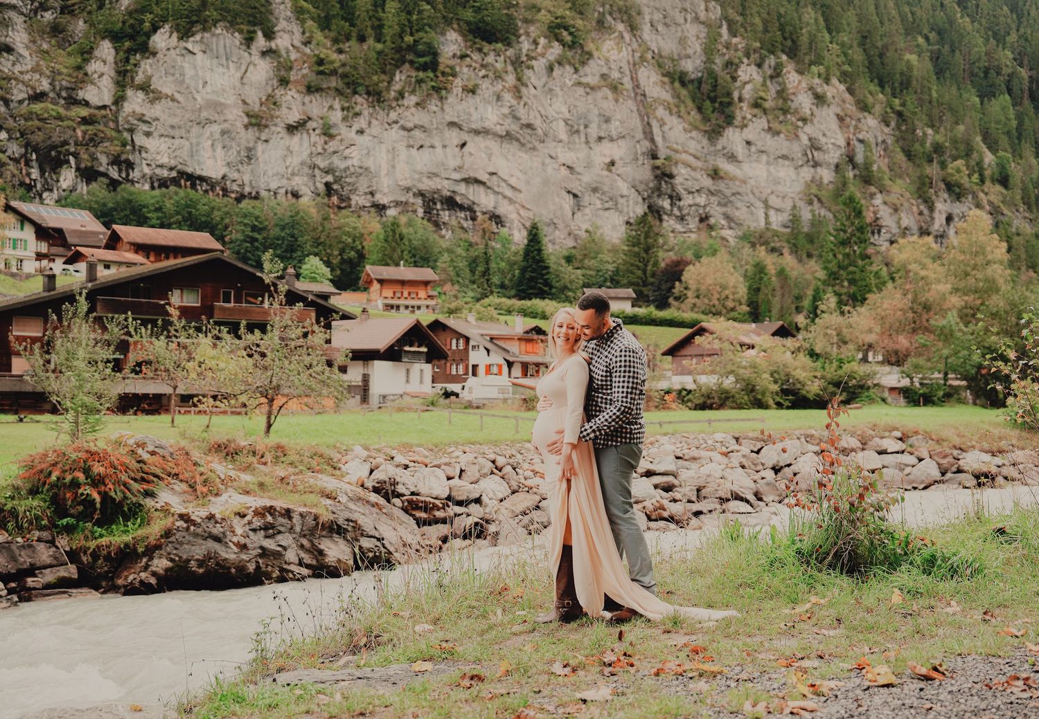Loving couple embracing during maternity photoshoot with charming Swiss alpine village and green mountains in background.