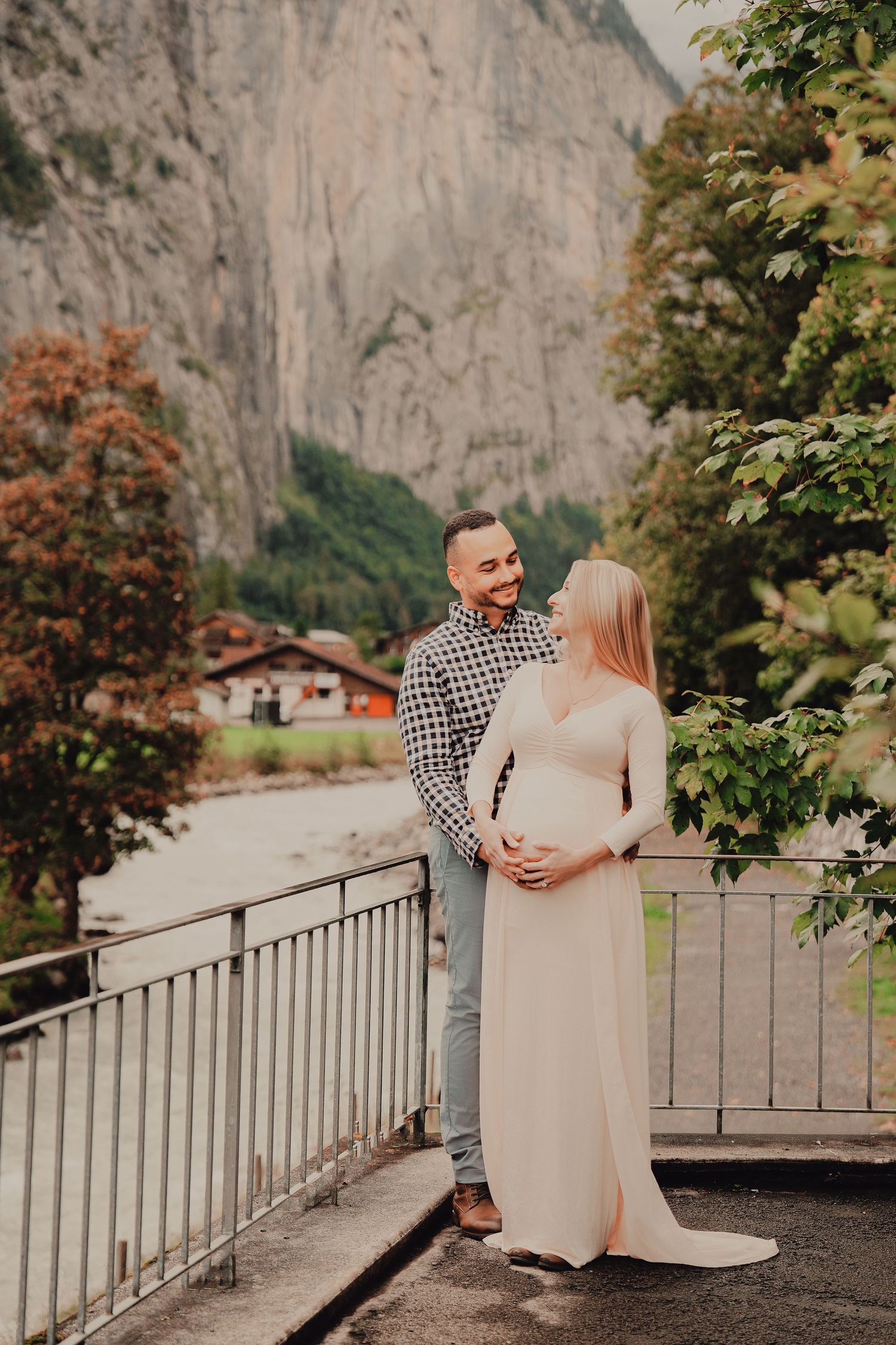 Couple embracing on bridge, pregnant woman in blush dress, surrounded by Swiss alpine scenery.