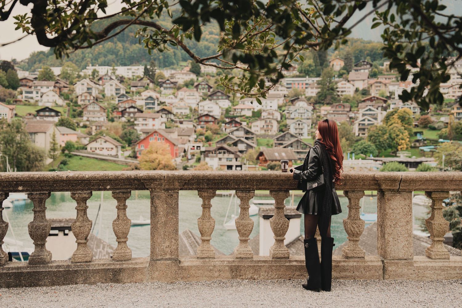 Stylish woman in black outfit admires colorful Swiss village from ornate stone balcony overlooking lake.