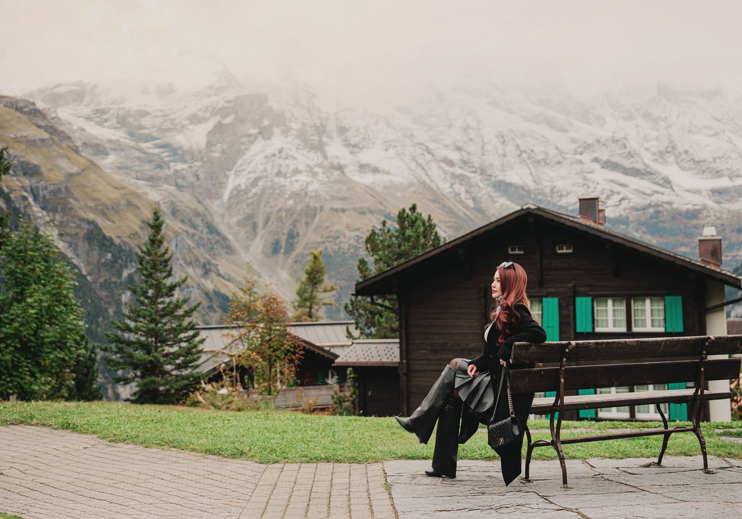 Woman with pink hair sits on a bench overlooking snow-capped Alps and a wooden chalet in Switzerland.