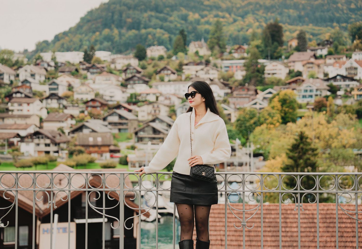 Woman in white sweater and black mini skirt poses on balcony overlooking Swiss alpine village.