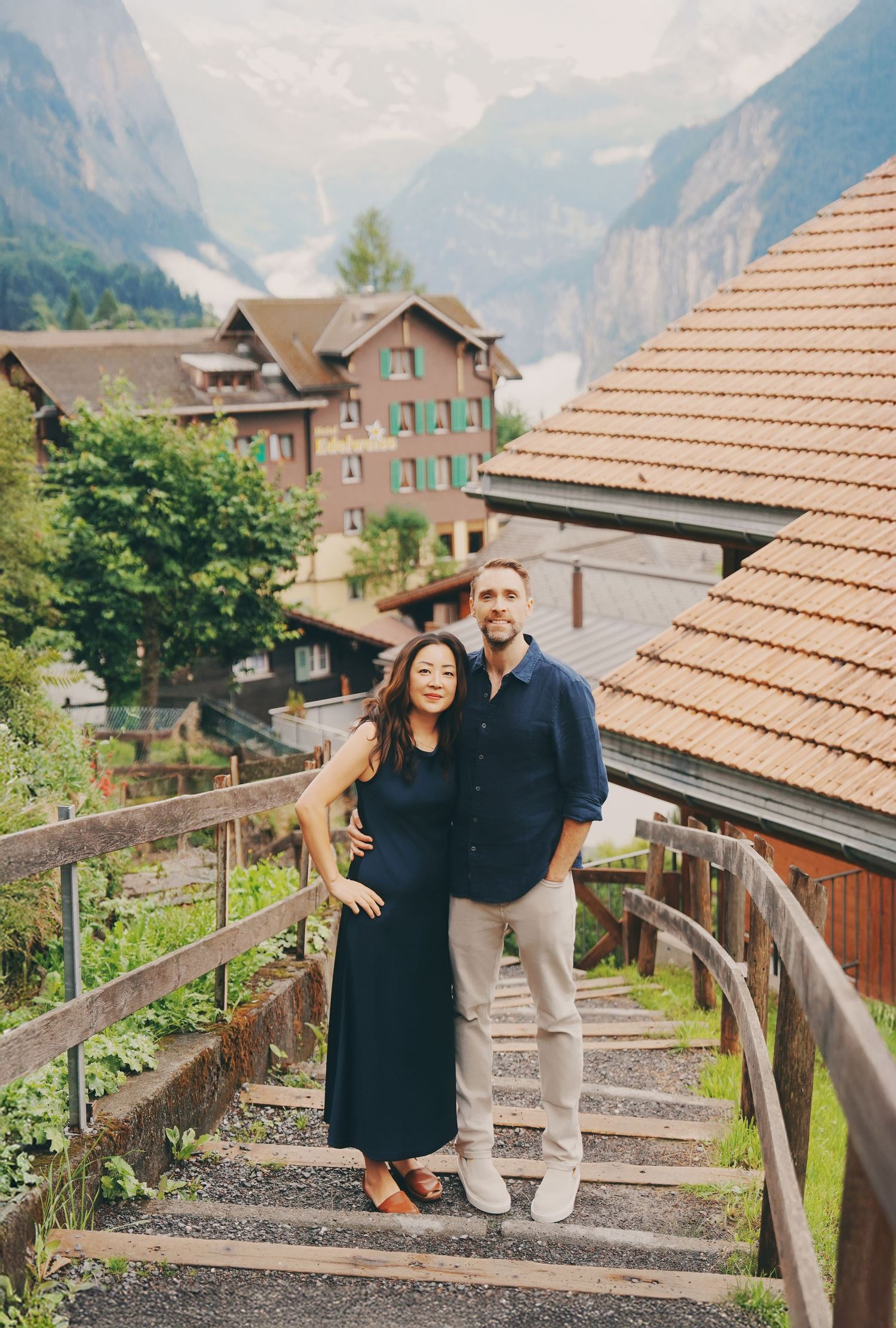 A couple in navy outfits poses on stone steps with a scenic Swiss alpine village behind them.