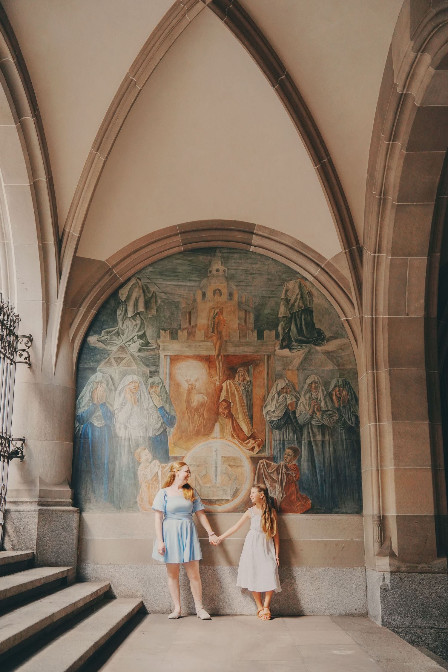 A woman and girl in blue and white dresses hold hands beneath Gothic arches and a colorful religious fresco.