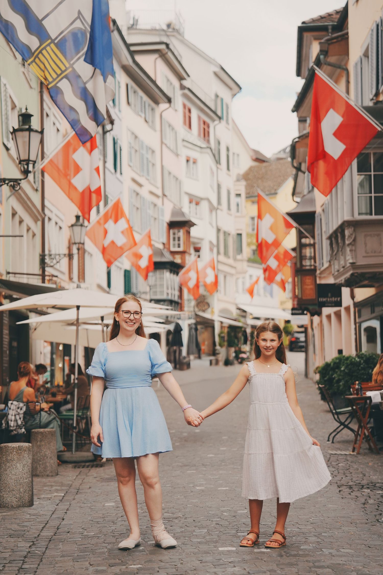 A woman and girl in matching blue and white dresses hold hands on a Swiss street decorated with flags.