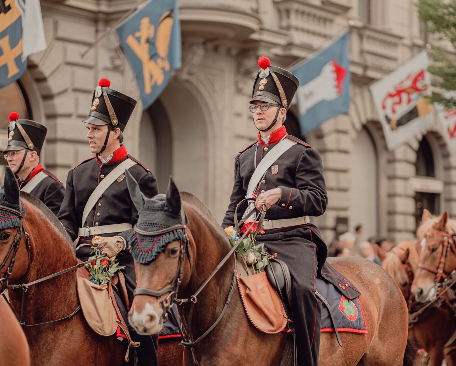Soldiers in historic black uniforms with red plumes ride horses in a traditional military parade.