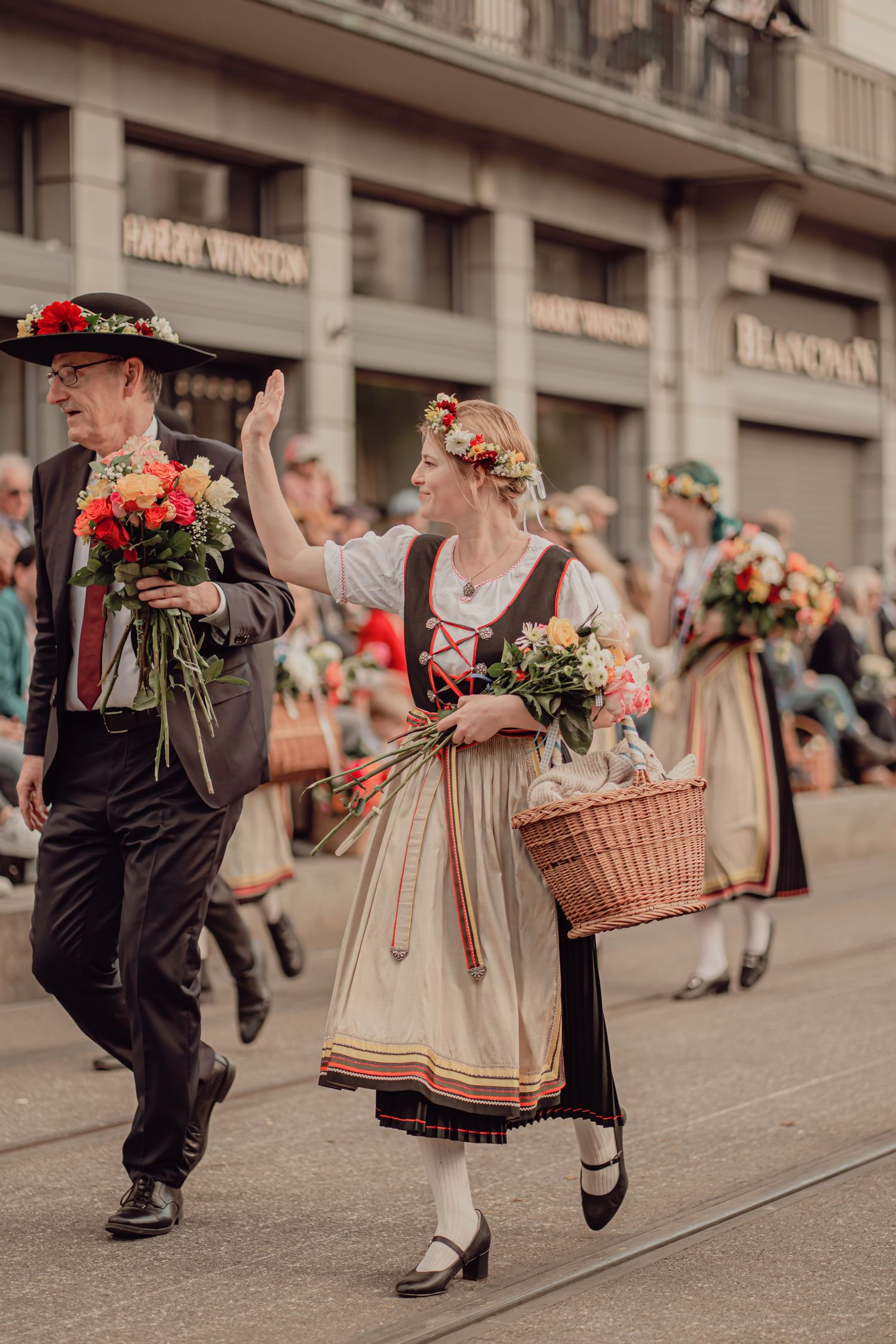 Man and woman in traditional Swiss folk costumes carry flower bouquets while marching in a parade.