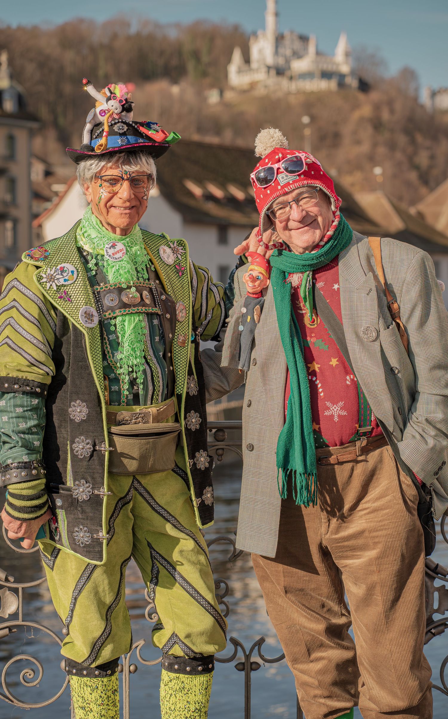 Two cheerful carnival revelers in vibrant costumes pose together on a bridge with a castle visible on the hillside behind them.