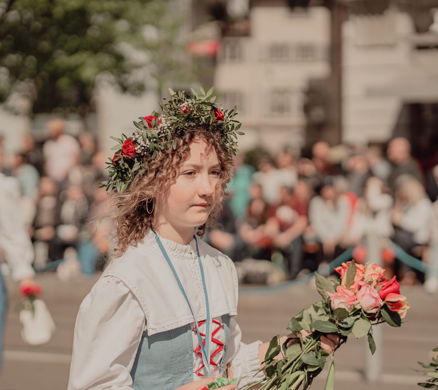 Young woman with floral crown holds roses in traditional folk costume at outdoor cultural parade.