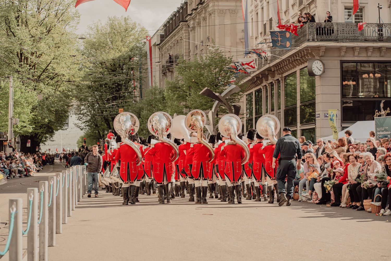 Marching band in red uniforms with large brass instruments perform for crowds lining a festive city street.