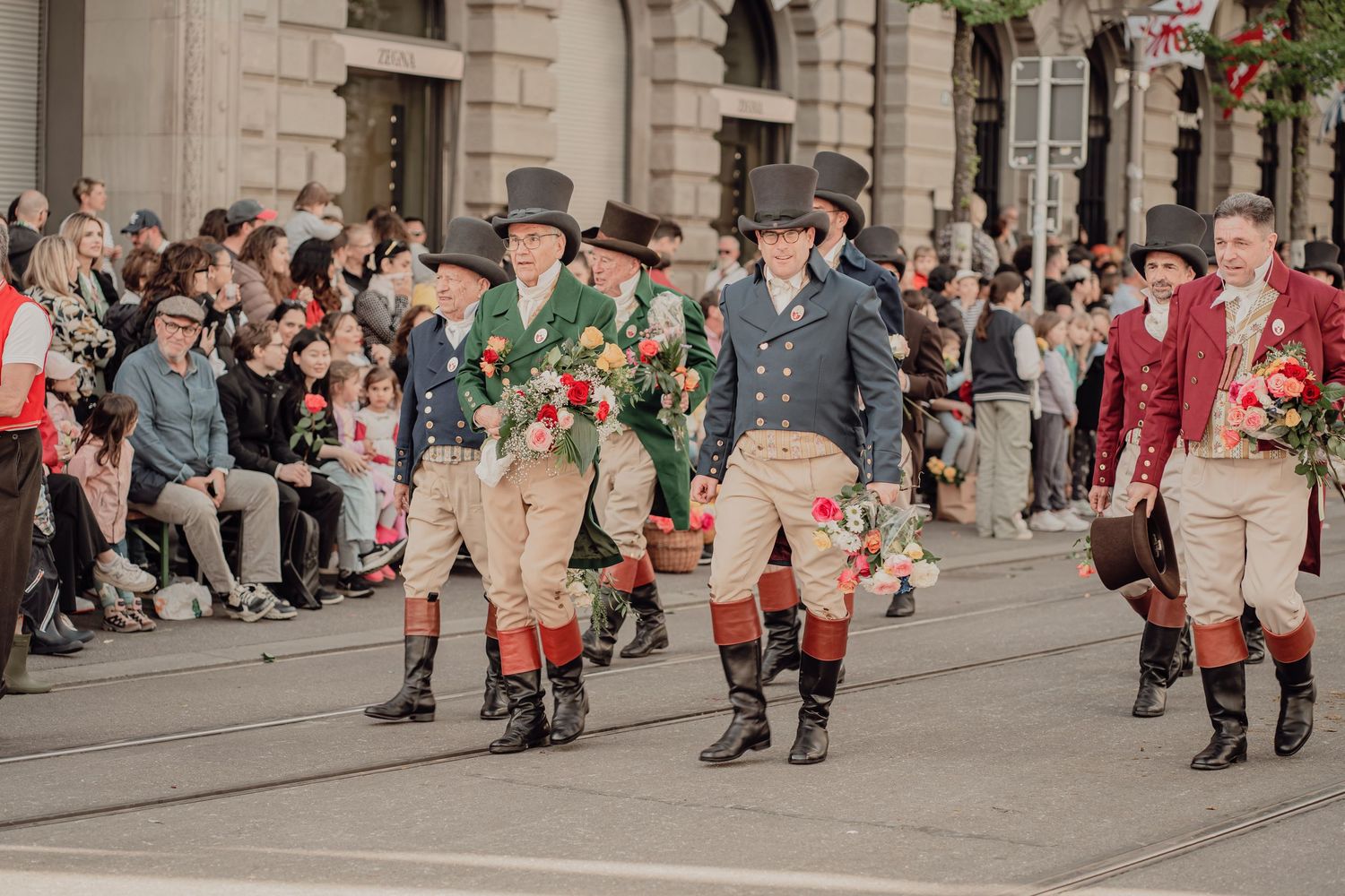 Men in historical costumes with black hats carry colorful flower bouquets in a traditional parade.