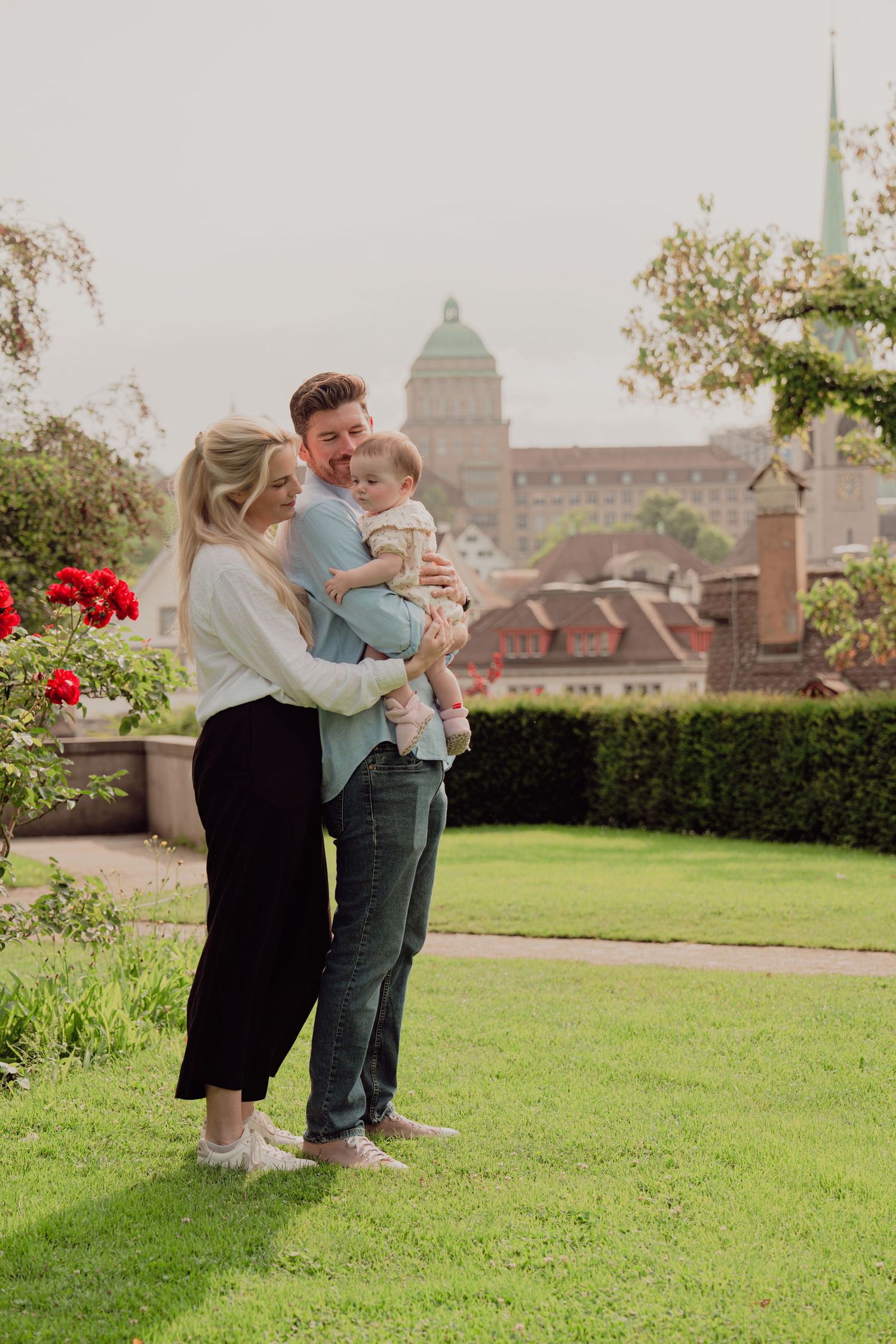 Happy family of three standing in lush garden with European city rooftops and dome visible.